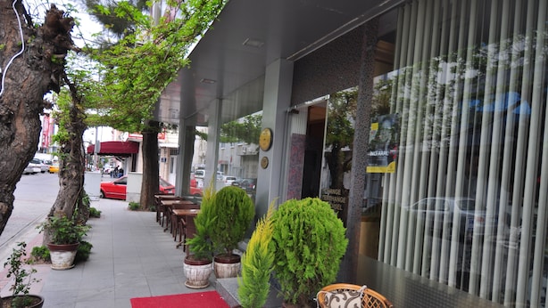 A sidewalk with potted plants, outdoor seating, and a red carpet leading to a building with vertical blinds. - Mini Fuar Hotel (Izmir)