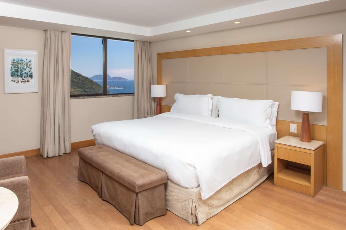 Ein Hotelzimmer mit einem großen Bett, Nachttischlampen, einer Bank, Blick auf das Meer und einem gerahmten Bild an der Wand.
