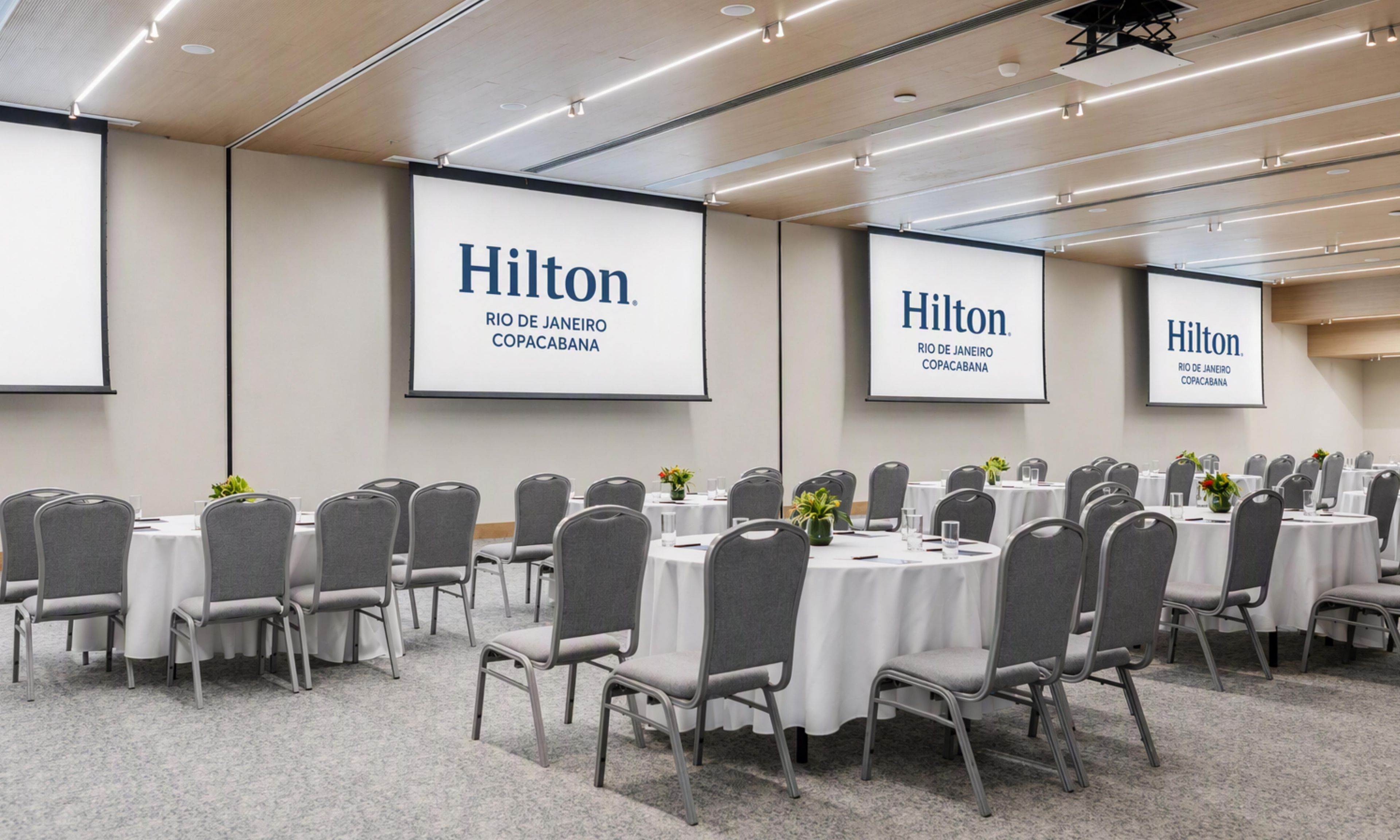 A conference room with multiple tables and chairs, large screens displaying the Hilton logo, and a carpeted floor.