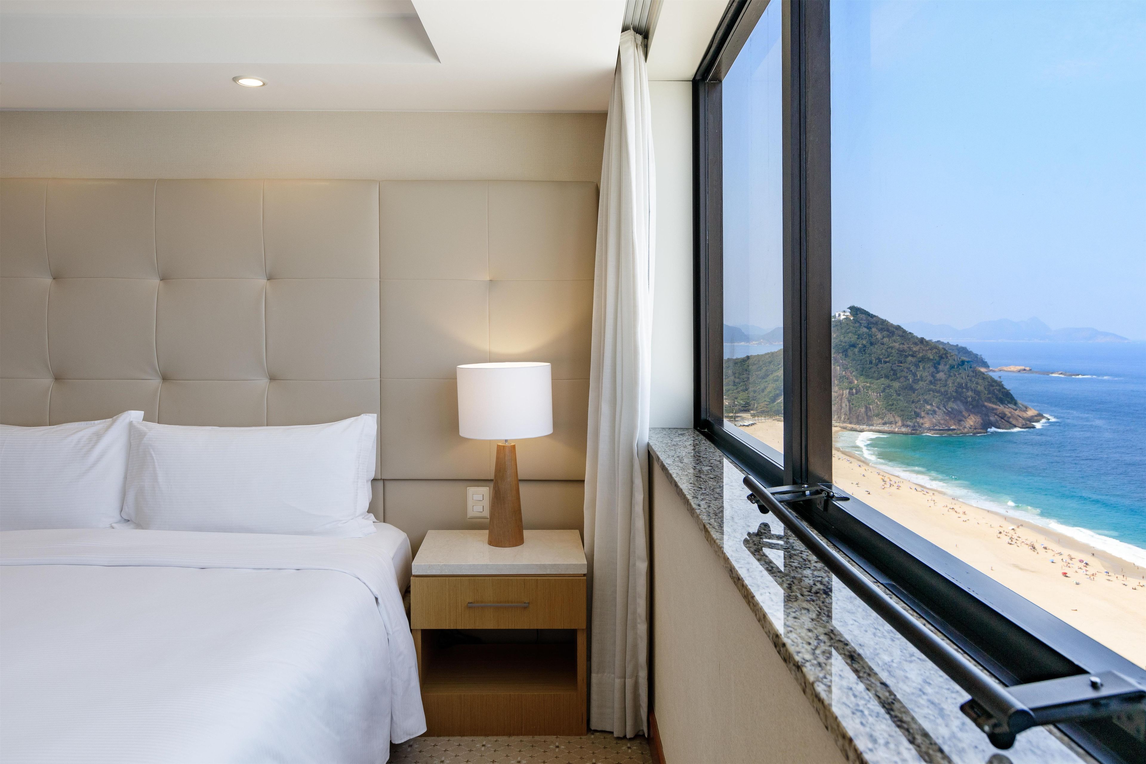 A hotel room with a bed, a nightstand, a lamp, a window with a view of a beach and mountains.