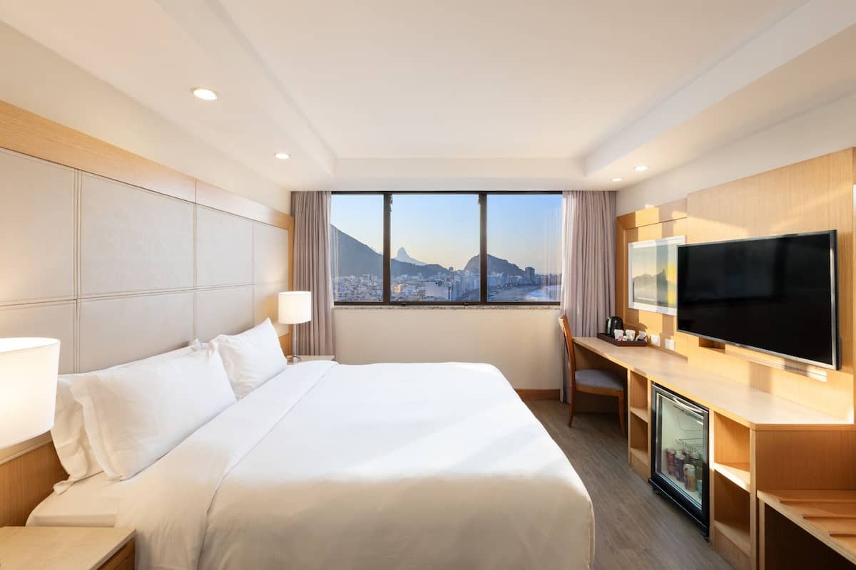 Ein Hotelzimmer mit einem großen Bett, einem Schreibtisch mit Fernseher und Blick auf eine Stadtlandschaft.