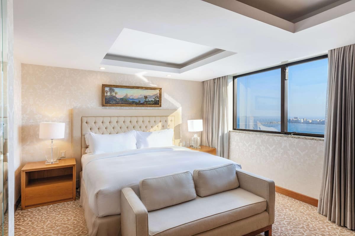 Ein Hotelzimmer mit einem großen Bett, einer Couch, einem Nachttisch mit Lampe, einem Bild an der Wand und Blick auf die Stadt durch große Fenster.