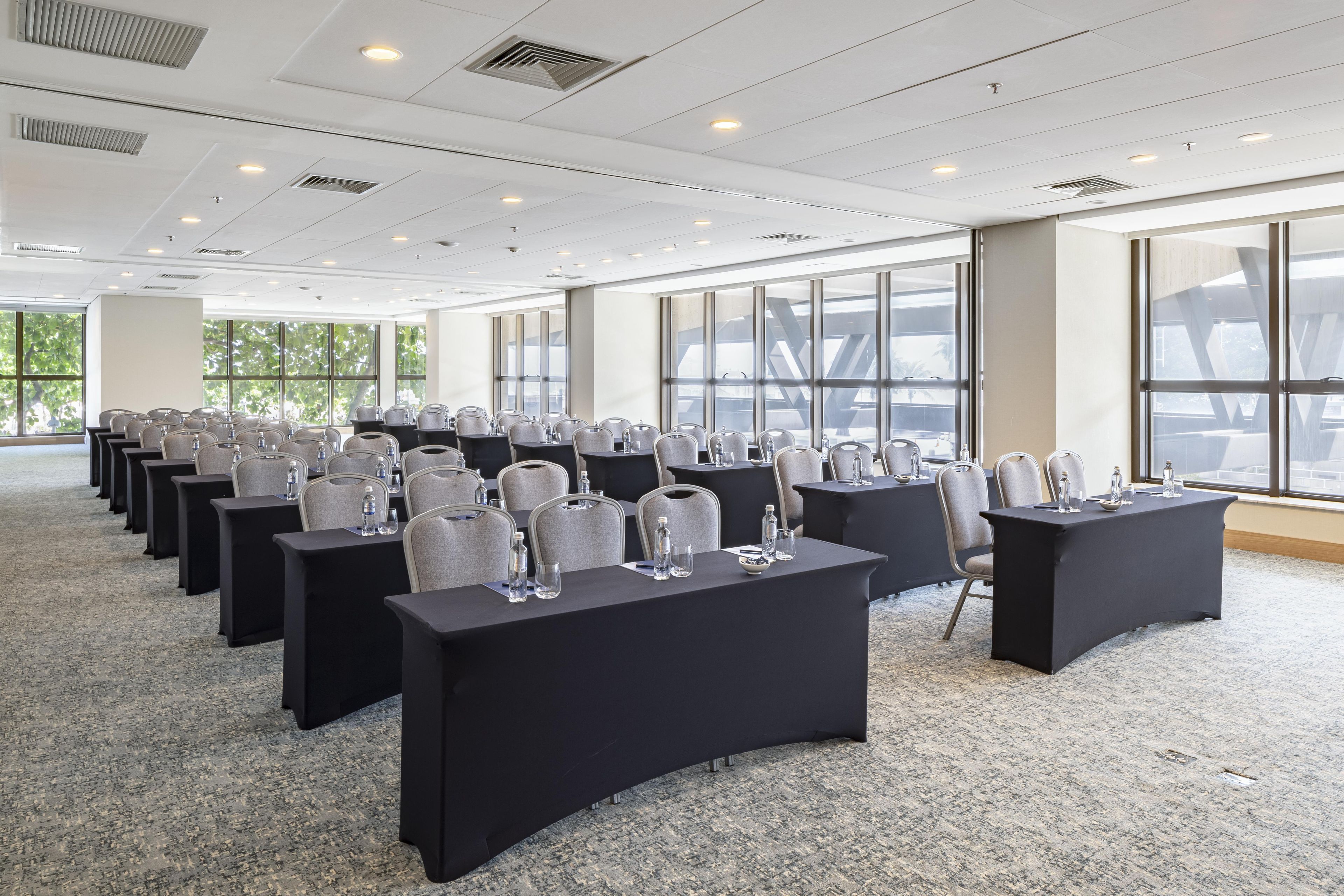 Un salón de conferencias con filas de mesas y sillas, ventanas amplias e iluminación empotrada en el techo.
