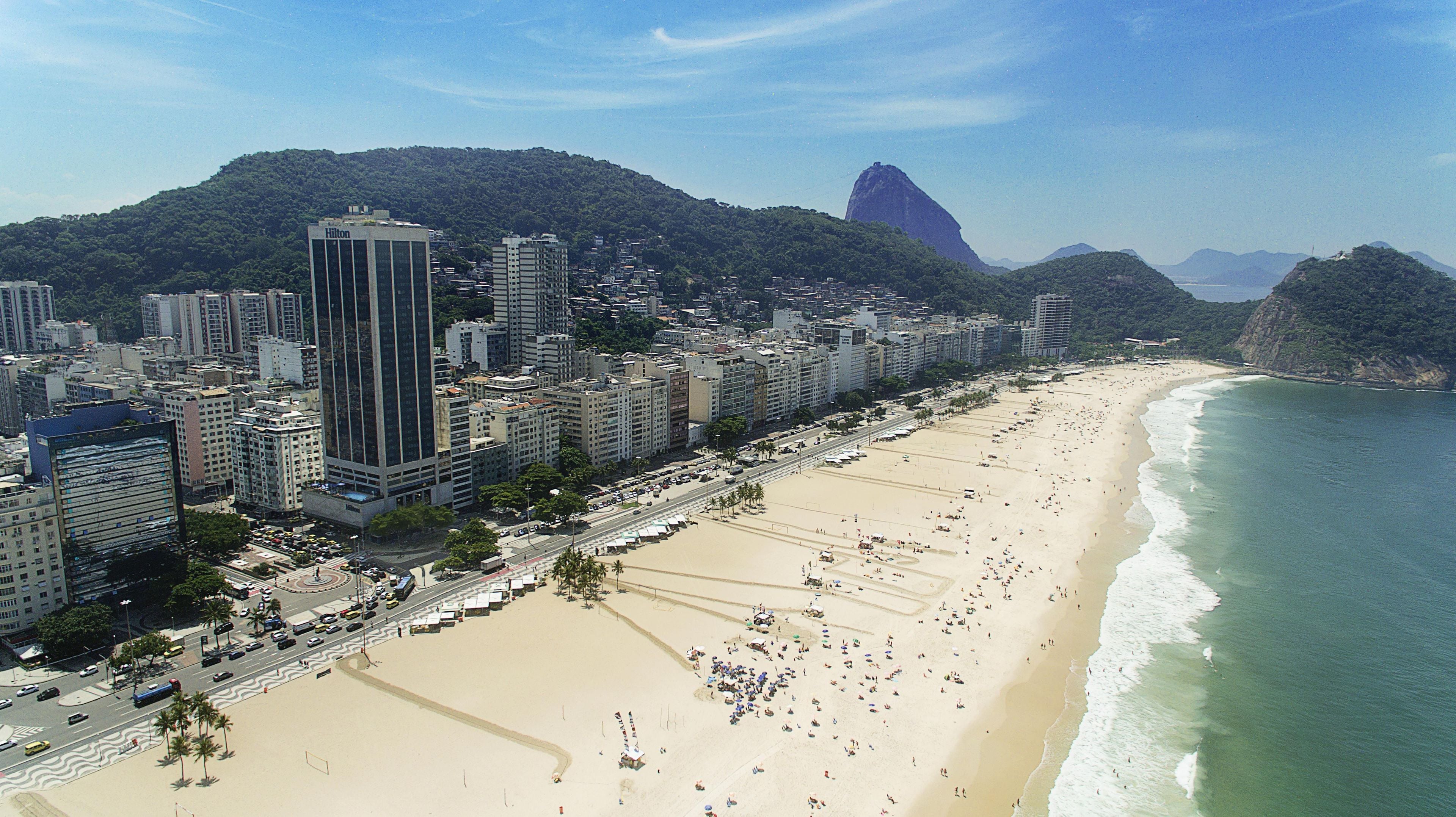 A coastal cityscape with a wide sandy beach, high-rise buildings, and a prominent mountain in the background.