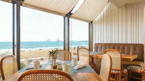 A beachside restaurant with a view of the ocean, wooden tables and chairs, and a cushioned seating area. - Hilton Copacabana Rio de Janeiro (Rio de Janeiro)