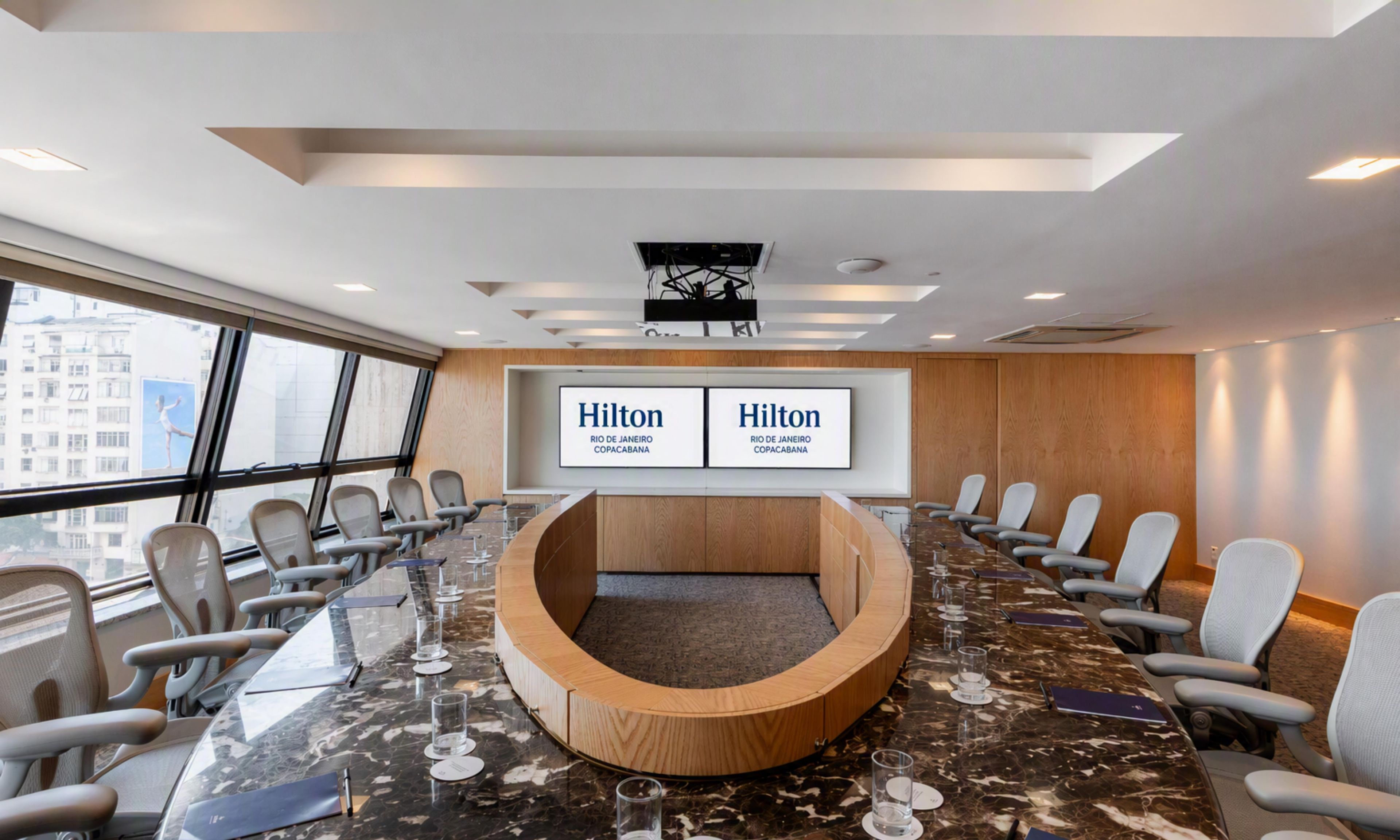 Un salón de conferencias con una larga mesa, varias sillas y una pantalla grande que muestra 'Hilton Rio de Janeiro Copacabana'.