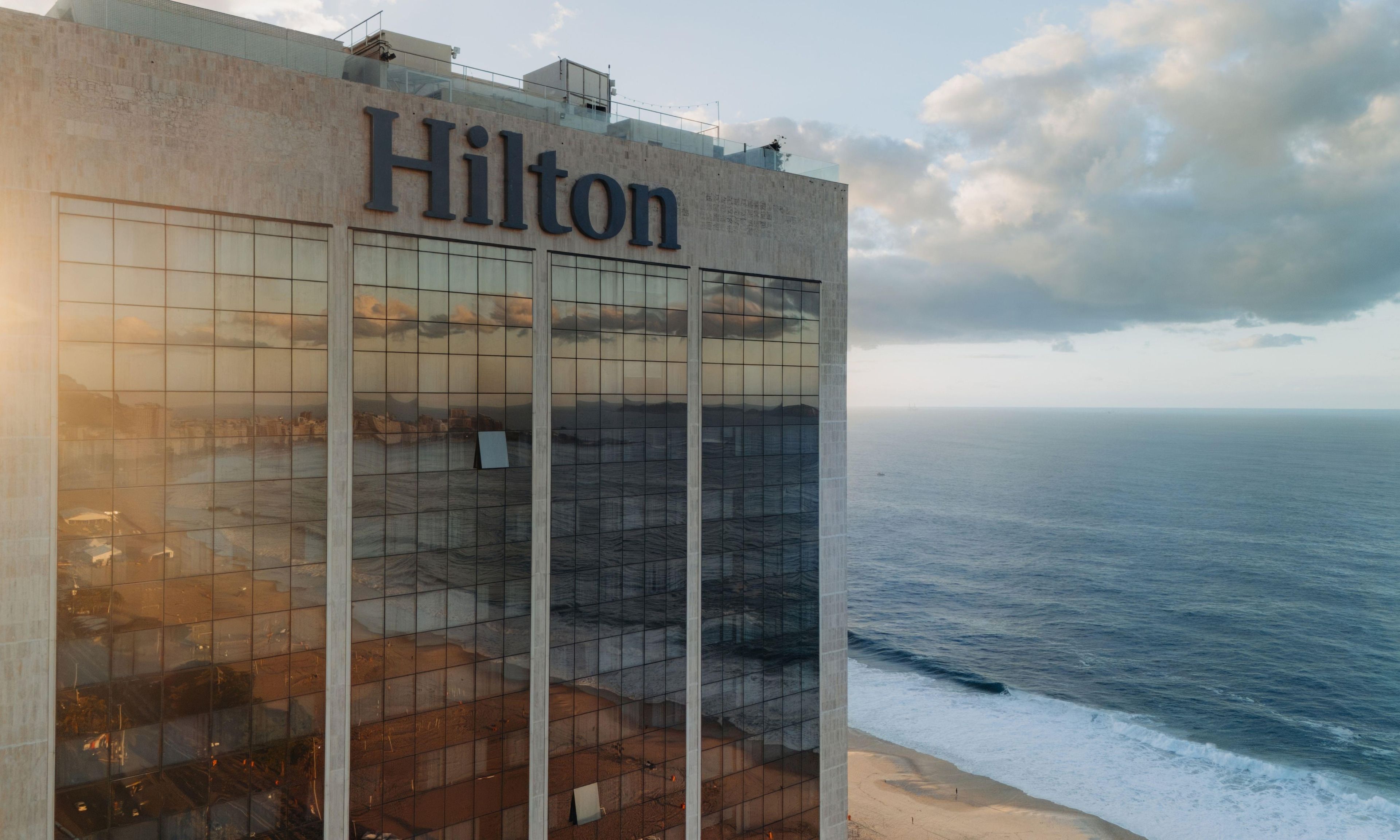 A Hilton hotel building with a large glass facade reflecting the surrounding cityscape and ocean view.