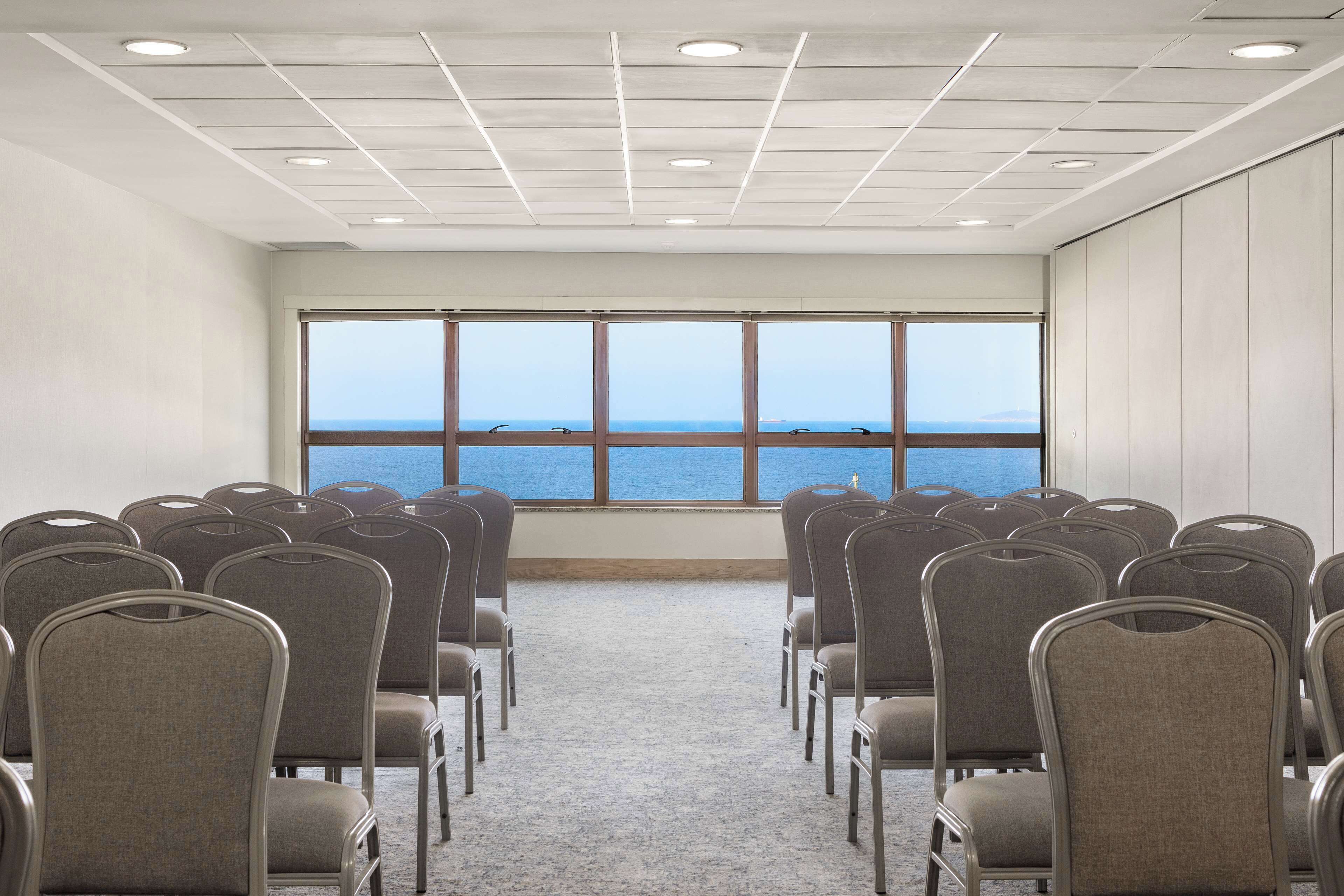 Un salón de conferencias con filas de sillas frente a un ventanal con vistas al mar.