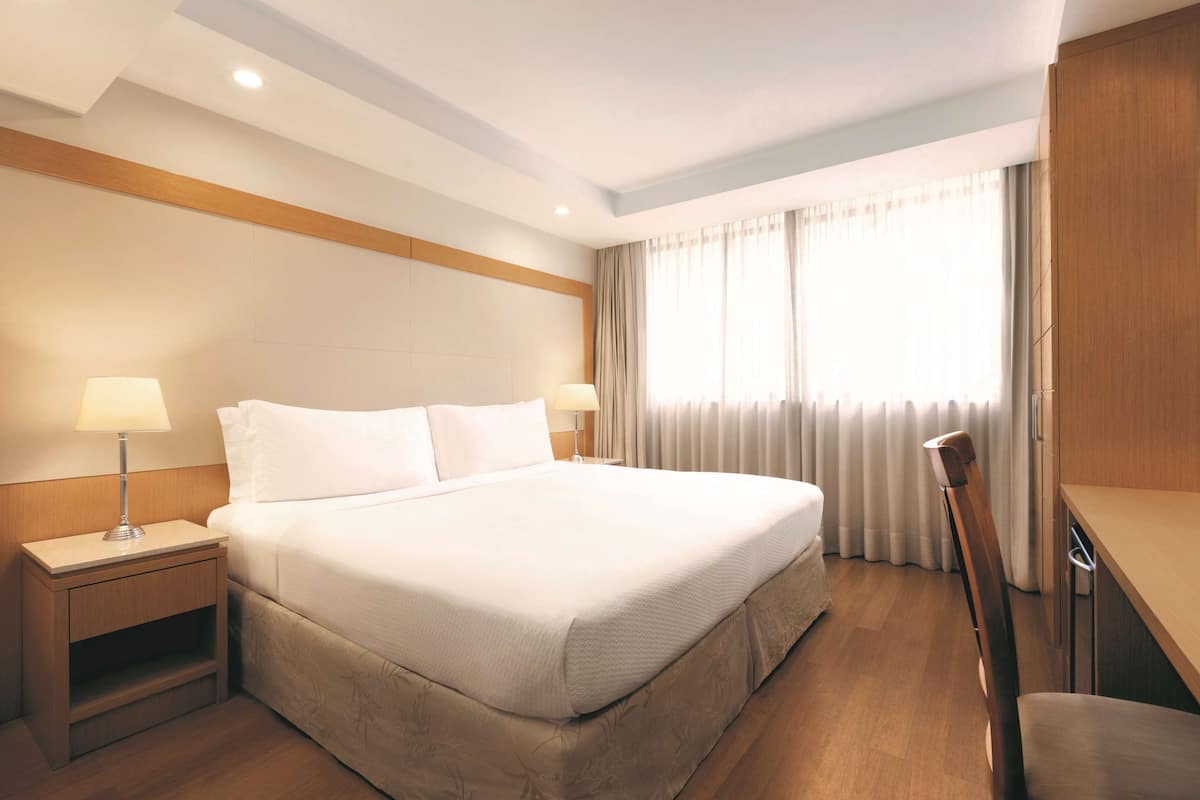 Ein Hotelzimmer mit einem großen Bett, zwei Nachttischen inklusive Lampen, einem Holz-Kopfteil, einem Schreibtisch mit Stuhl sowie einem Fenster mit Vorhängen.