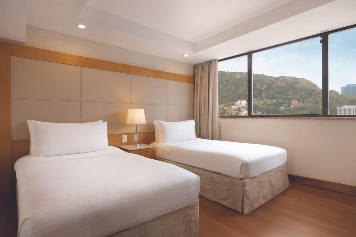 Ein Hotelzimmer mit zwei Betten, einem Nachttisch mit Lampe, einem großen Fenster und Blick auf eine Hügel- und Stadtlandschaft.