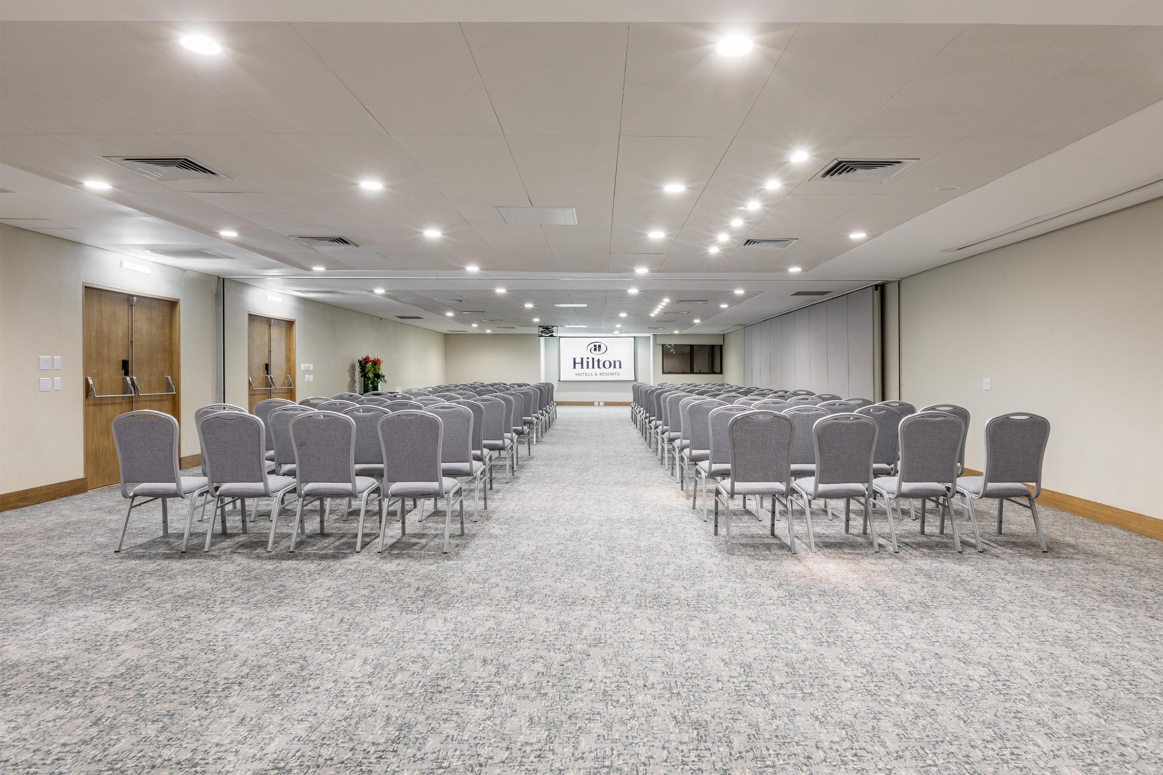 A hotel conference room with rows of grey chairs, carpeted floor, wooden doors, and a sign for Hilton Hotels & Resorts.