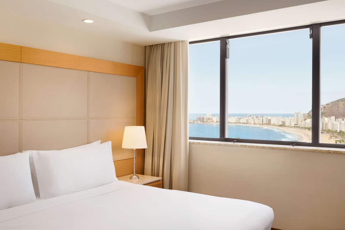 Ein Hotelzimmer mit Bett, Nachttisch, Lampe und einem großen Fenster mit Blick auf Stadt und Strand.