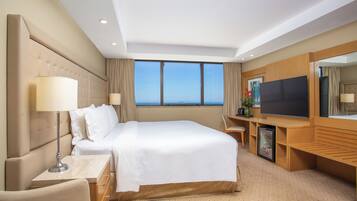 A hotel room with a large bed, a flat-screen TV, a minibar, and a view of the ocean.