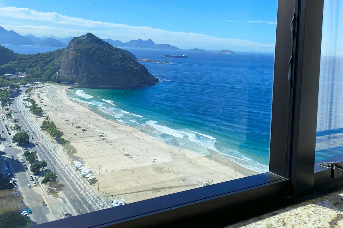 Blick vom Fenster auf die Küste, mit Strand, einer parallel zur Küste verlaufenden Straße, einer Fahne auf einem Hügel und fernen Bergen.