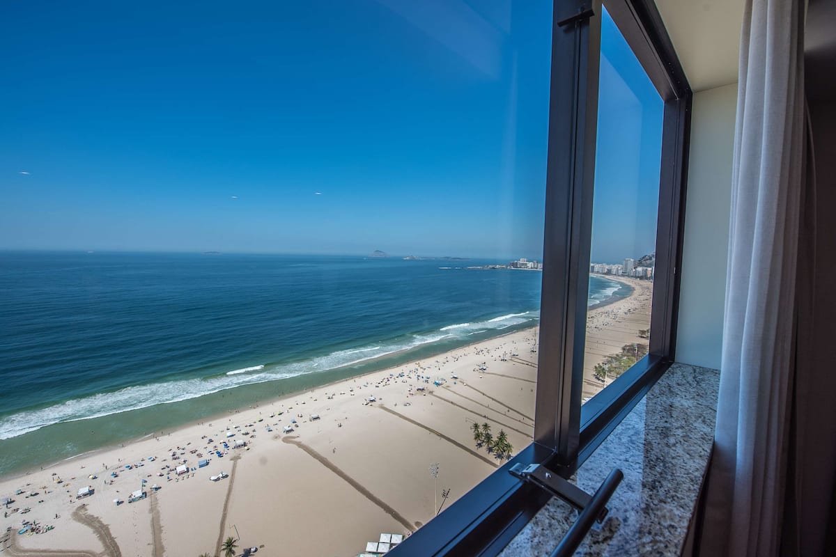 Blick auf einen Sandstrand aus einem Fenster, mit Sonnenschirmen und Menschen.