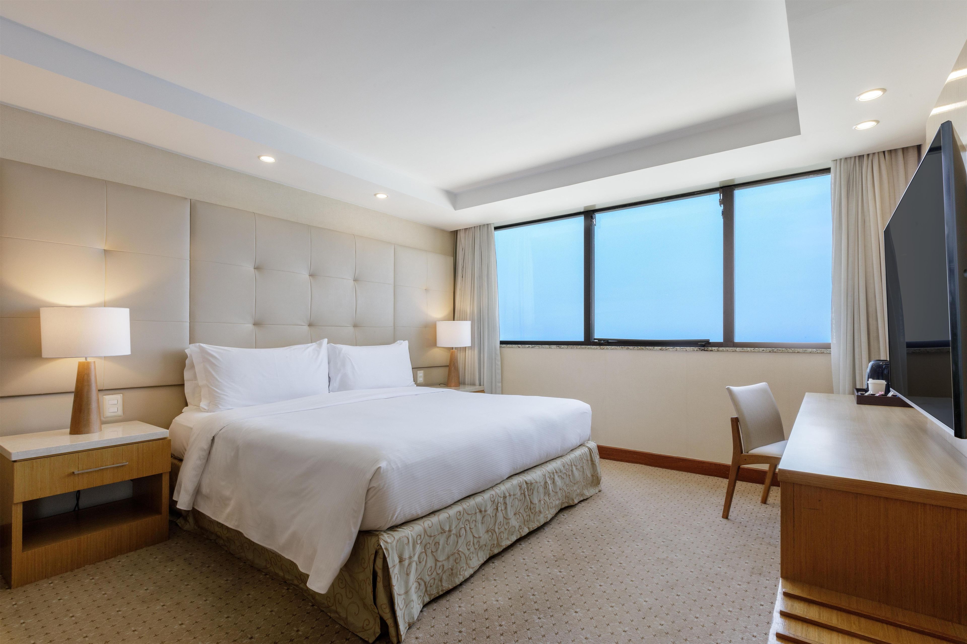 Quarto de hotel com uma cama grande, luminárias de cabeceira, uma escrivaninha e uma televisão.