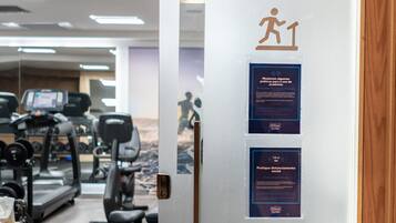 Un gimnasio con cintas de correr, pesas y equipo de ejercicio. Hay carteles en la puerta sobre distanciamiento social y medidas de higiene.