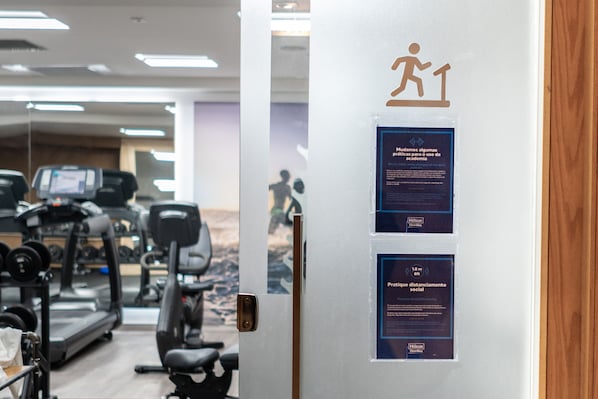 A gym with treadmills, weights, and exercise equipment. There are signs on the door about social distancing and hygiene practices. - Hilton Copacabana Rio de Janeiro (Rio de Janeiro)