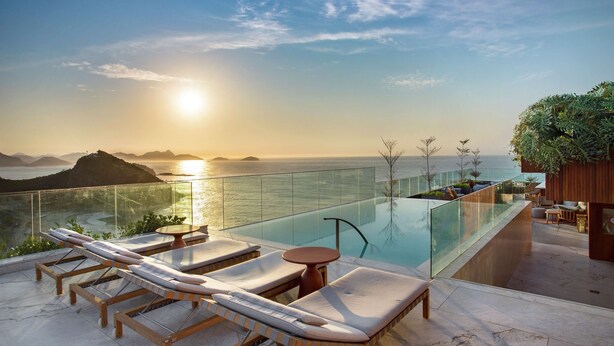 A rooftop infinity pool with lounge chairs and a table, overlooking a body of water and distant mountains during sunset. - Hilton Copacabana Rio de Janeiro (Rio de Janeiro)