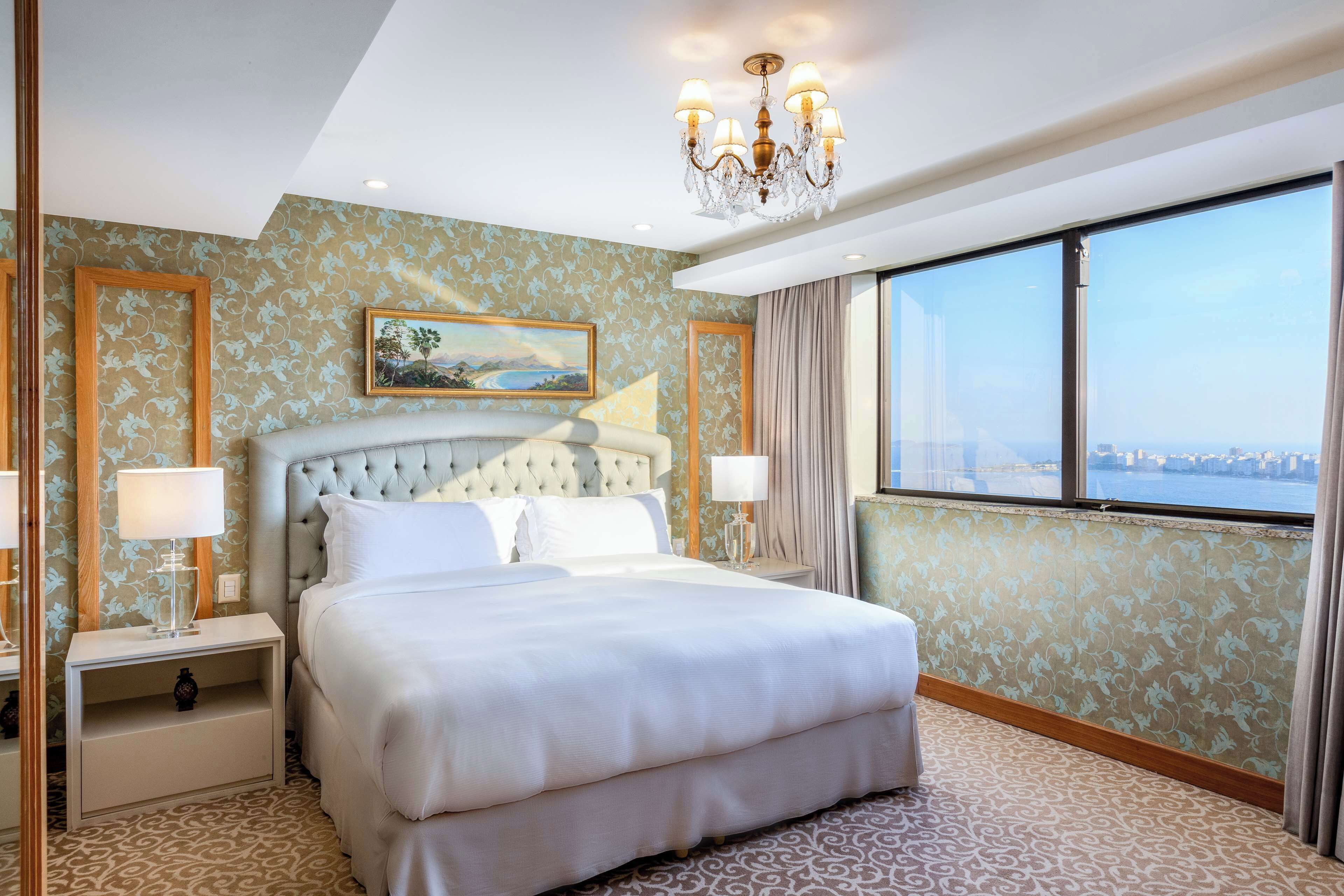 A spacious bedroom with a large bed, a chandelier, a nightstand, and a view of the city through large windows.