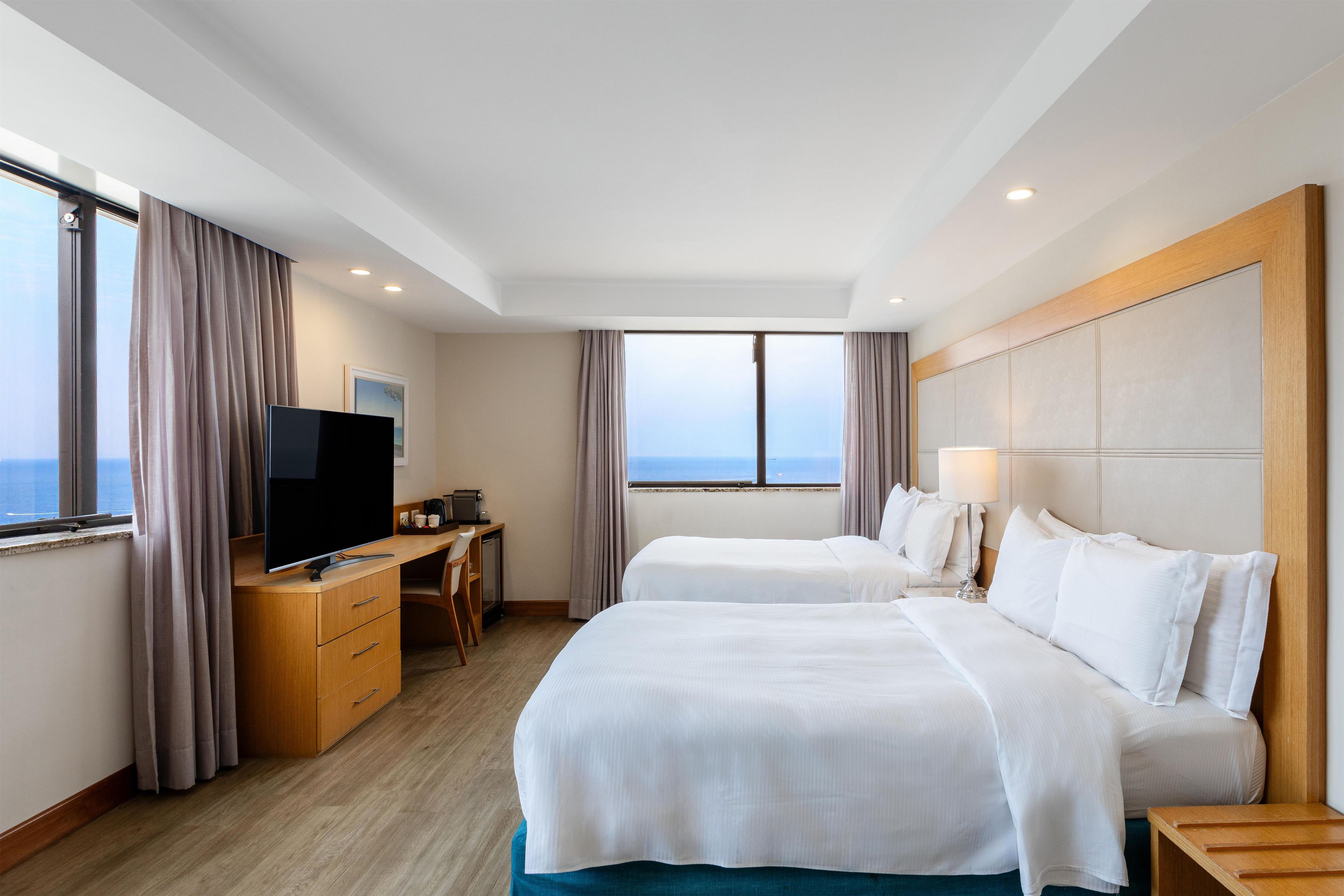 A hotel room with two beds, a desk, a television, and a view of the sea.