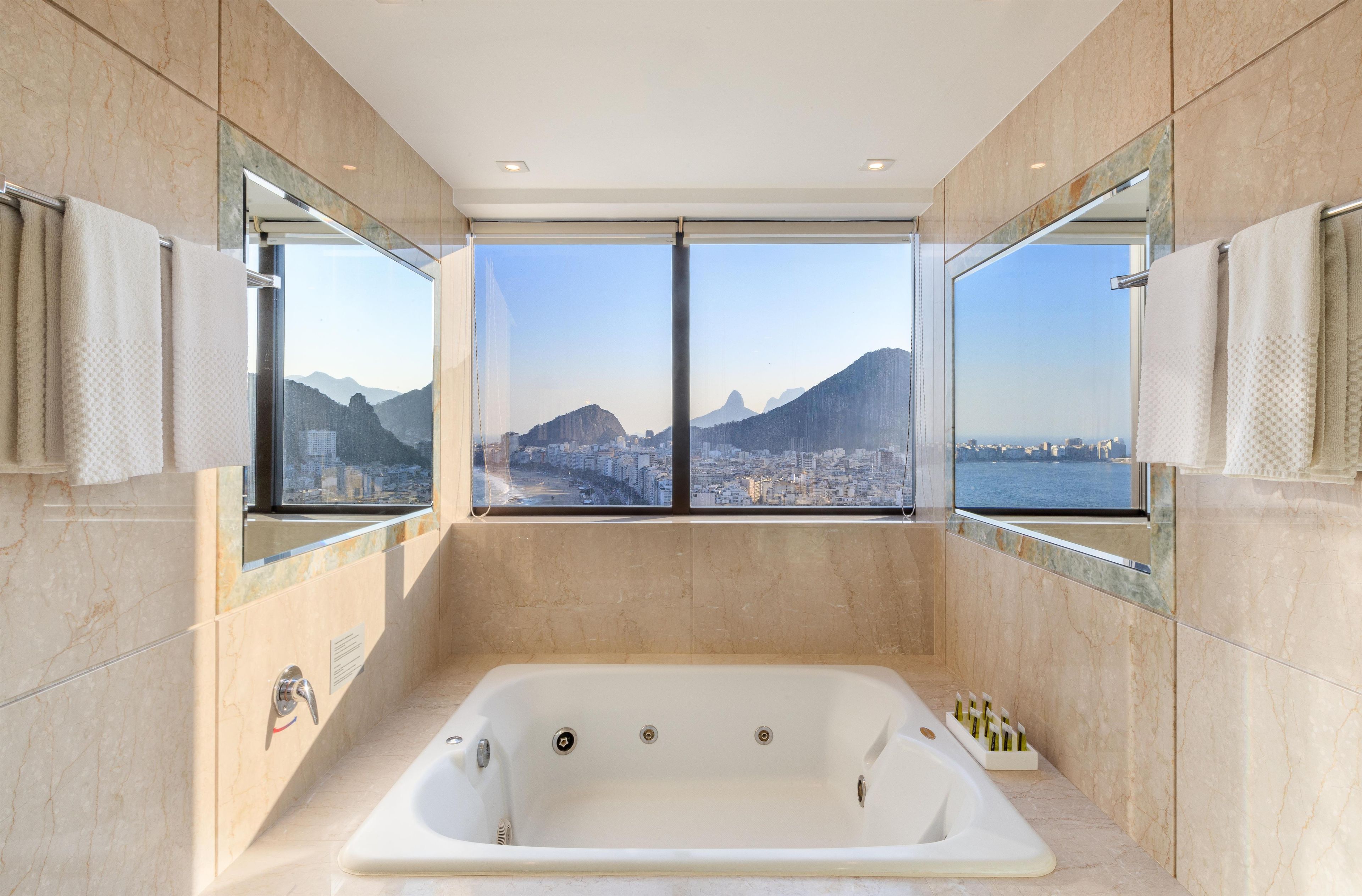 A modern bathroom with a large bathtub, two mirrors, and a city view.