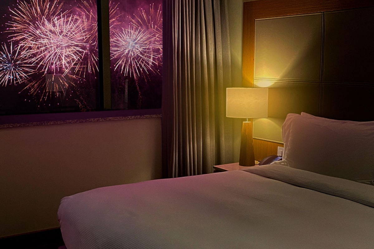 A hotel room with a bed, a nightstand, a lamp, and a window with fireworks visible outside.