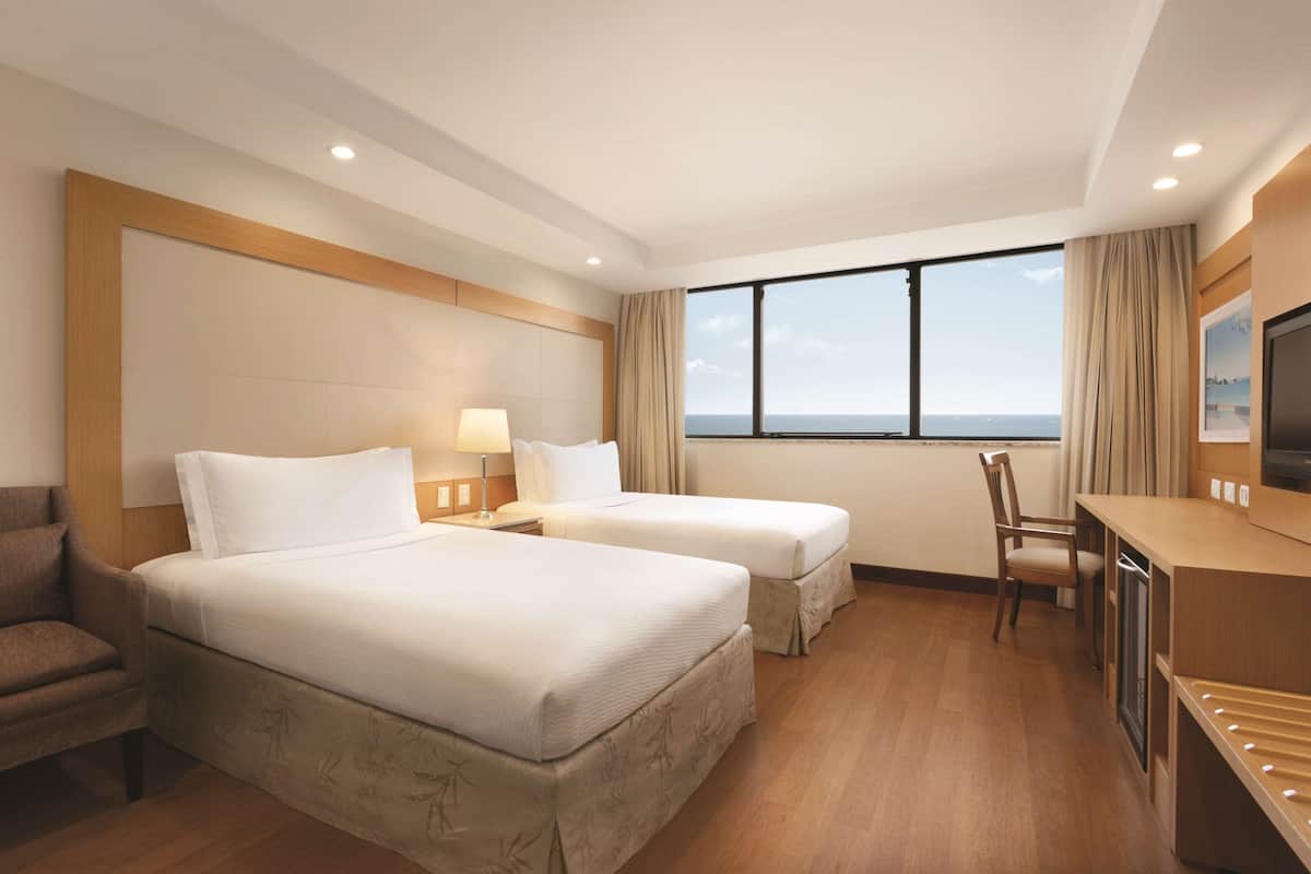 Ein Hotelzimmer mit zwei Betten, einem Schreibtisch, einem Stuhl, einem Fernseher und einem großen Fenster mit Meerblick.