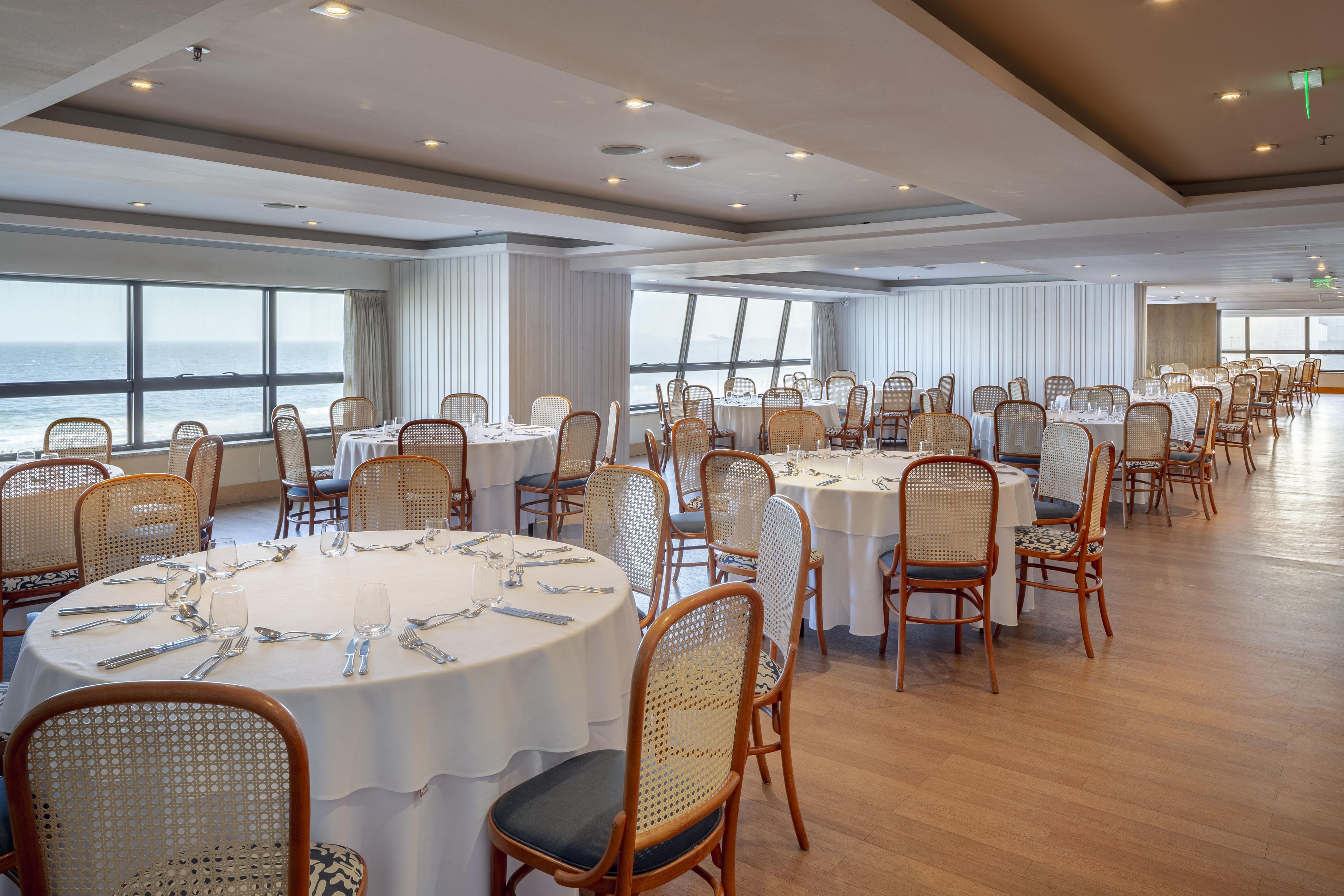 A spacious dining area with round tables and wicker chairs, large windows offering a view of the sea, and a modern ceiling design.
