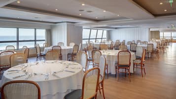 A spacious dining area with round tables and wicker chairs, large windows offering a view of the sea, and a modern ceiling design.