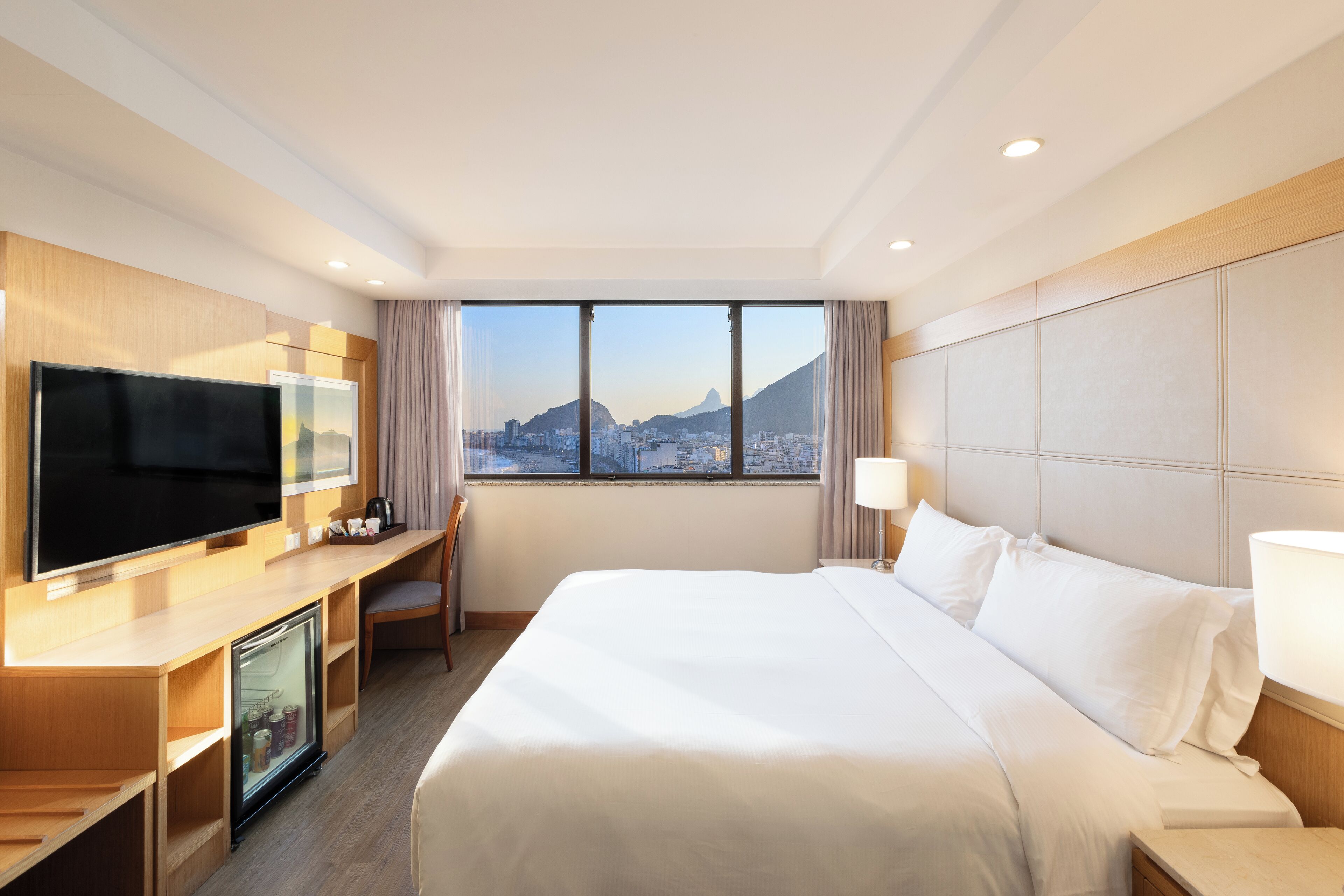 A hotel room with a bed, a TV, a desk with a refrigerator, and a view of a cityscape.