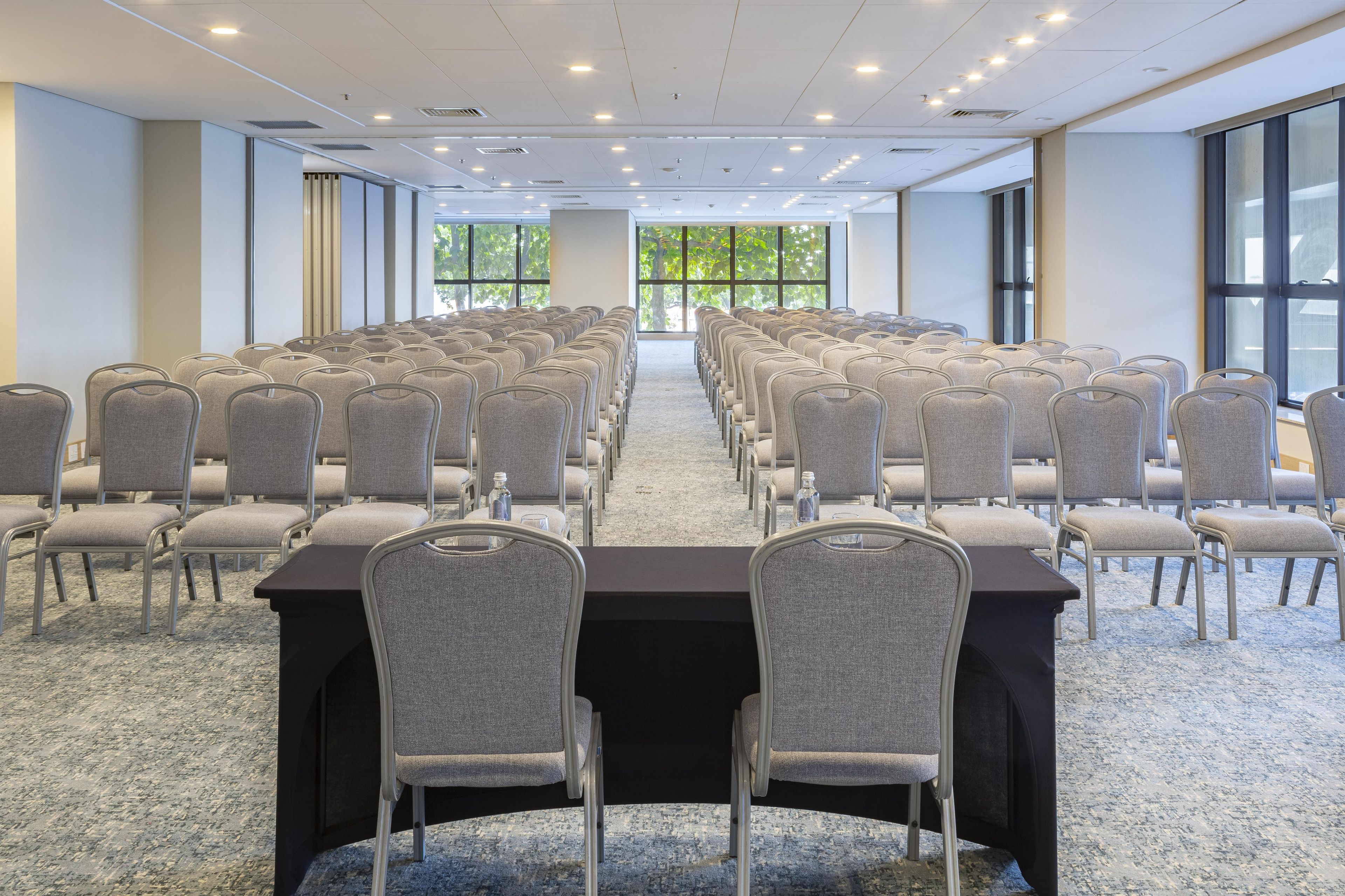 Sala de conferências com fileiras de cadeiras e mesas, janelas amplas e piso coberto de carpete.
