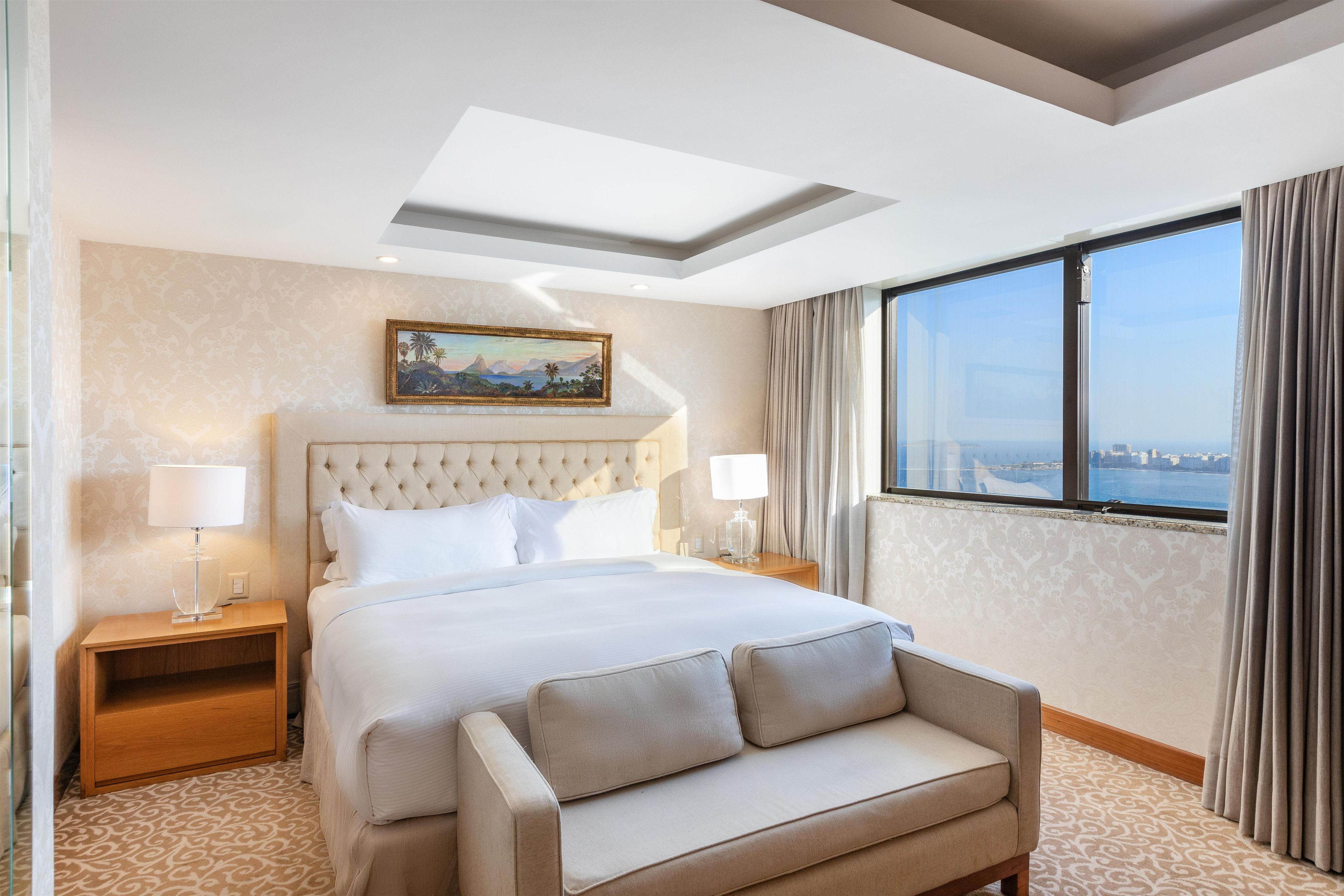 Quarto de hotel com cama, sofá, pintura, mesa de cabeceira, luminária e vista da cidade por uma grande janela.