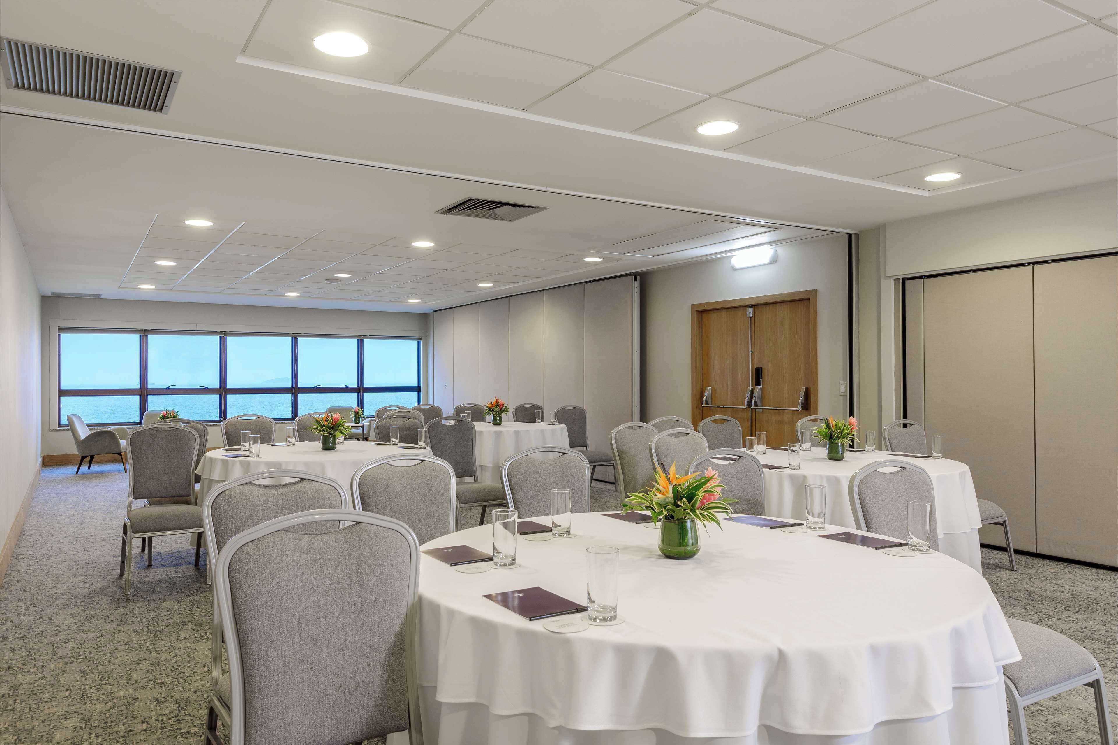 Un salón de conferencias con mesas redondas, sillas grises y manteles blancos. Hay jarrones con flores sobre las mesas, y la sala cuenta con grandes ventanales con vista al mar.