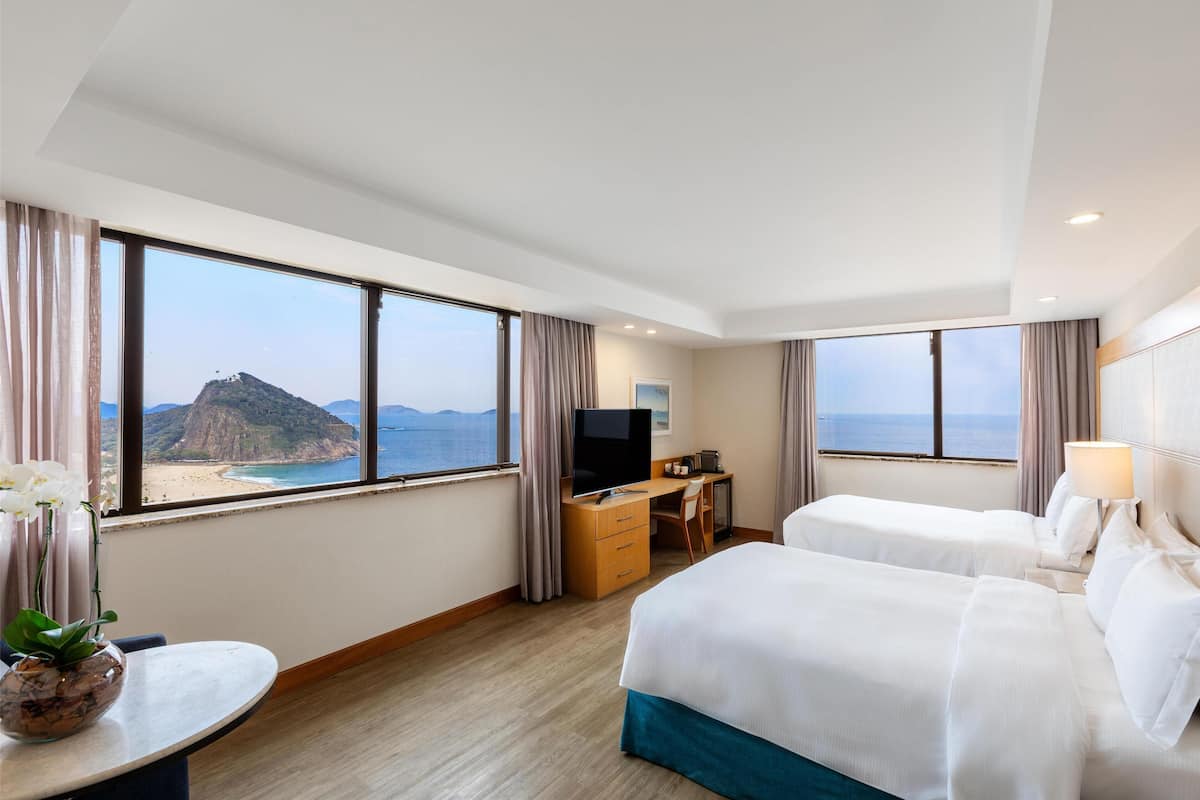 Ein Hotelzimmer mit zwei Betten, einem Schreibtisch, einem Fernseher und Blick auf Strand und Meer.