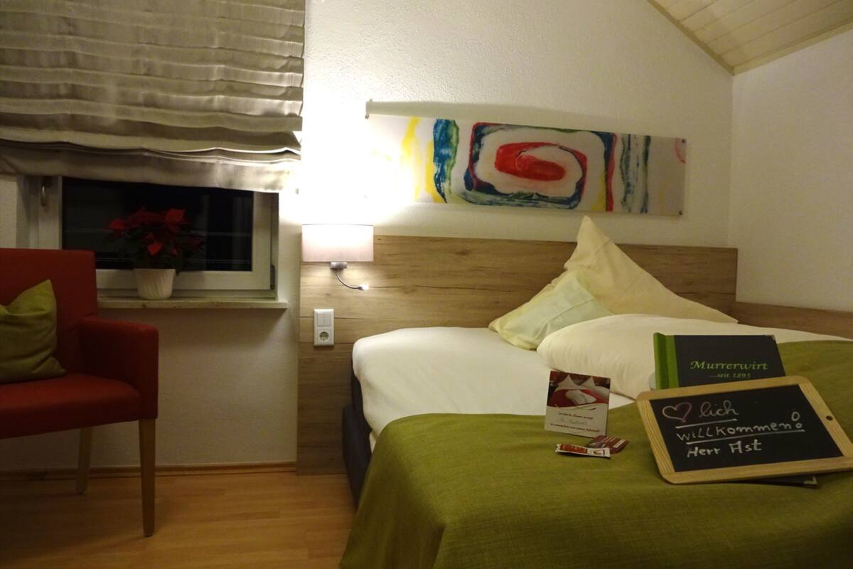 A hotel room with a wooden headboard, a bedside table with a lamp, a window with a view of the outside, and a bed with a green bedspread.