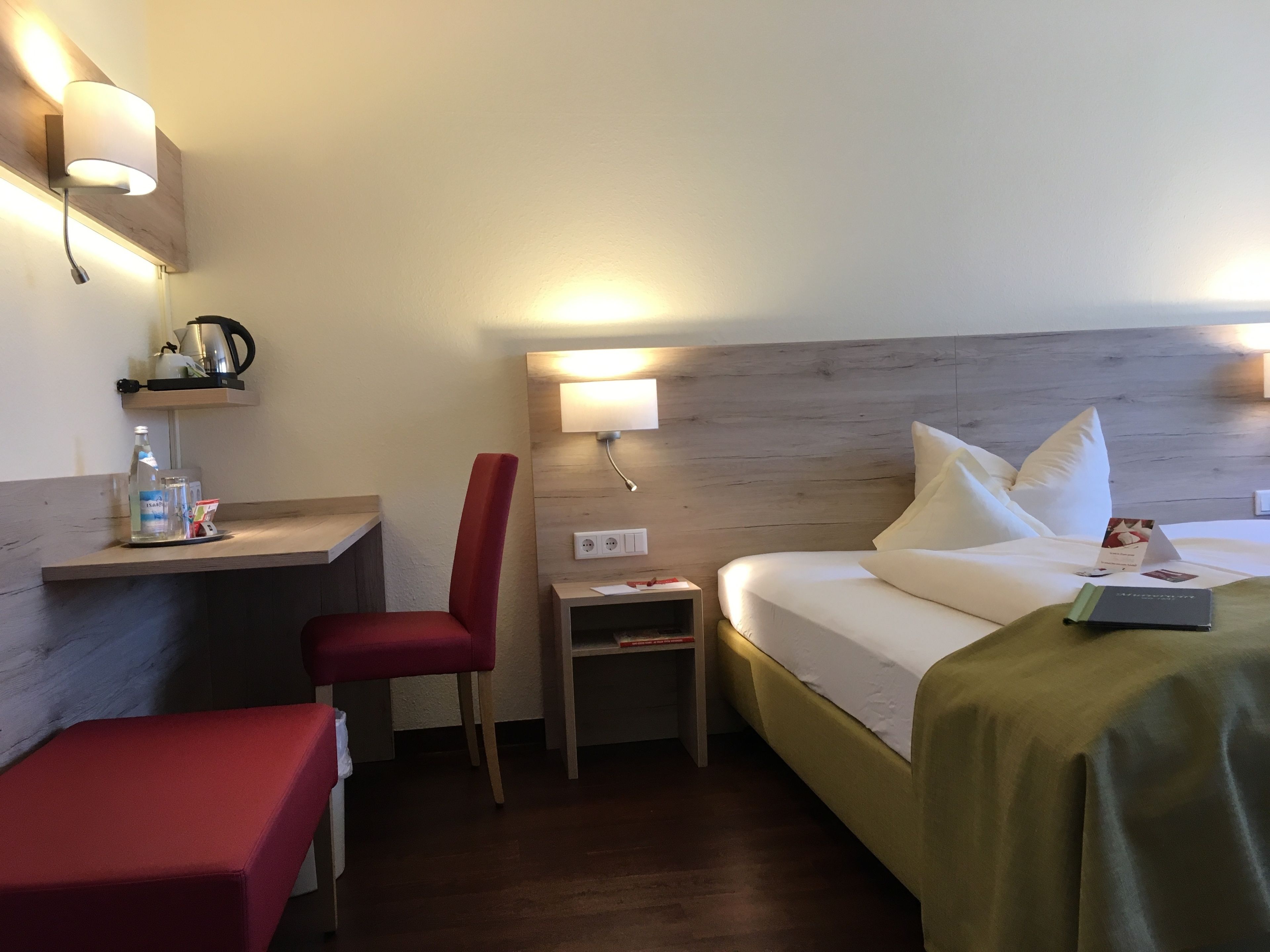 Une chambre d’hôtel comprenant un lit, un bureau, une chaise, une table de chevet et une étagère avec une bouilloire et une bouteille.