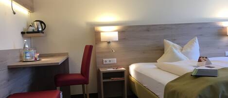 Une chambre d’hôtel comprenant un lit, un bureau, une chaise, une table de chevet et une étagère avec une bouilloire et une bouteille.
