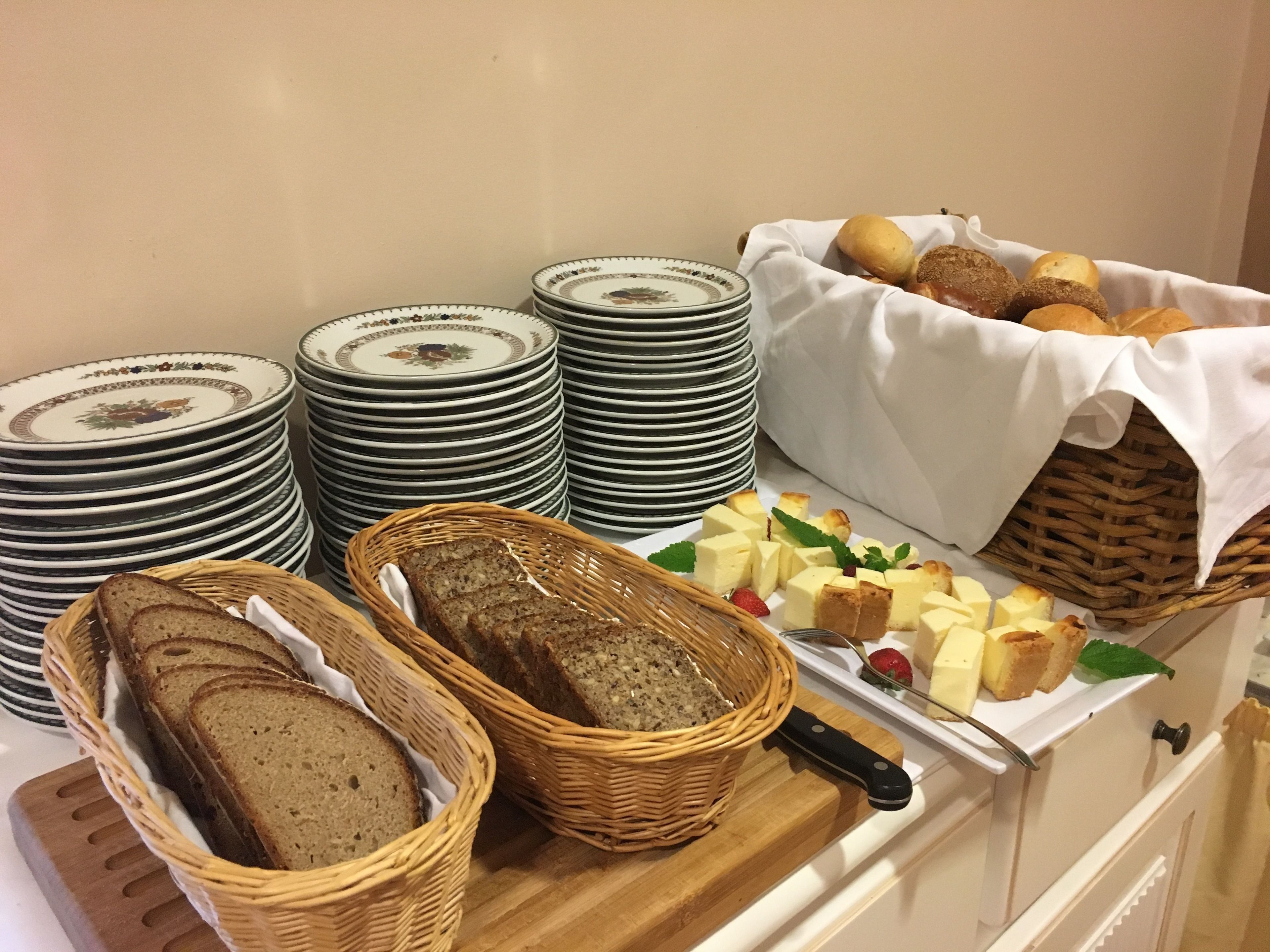A buffet table with stacks of plates, baskets of bread, and a variety of cheeses and pastries.