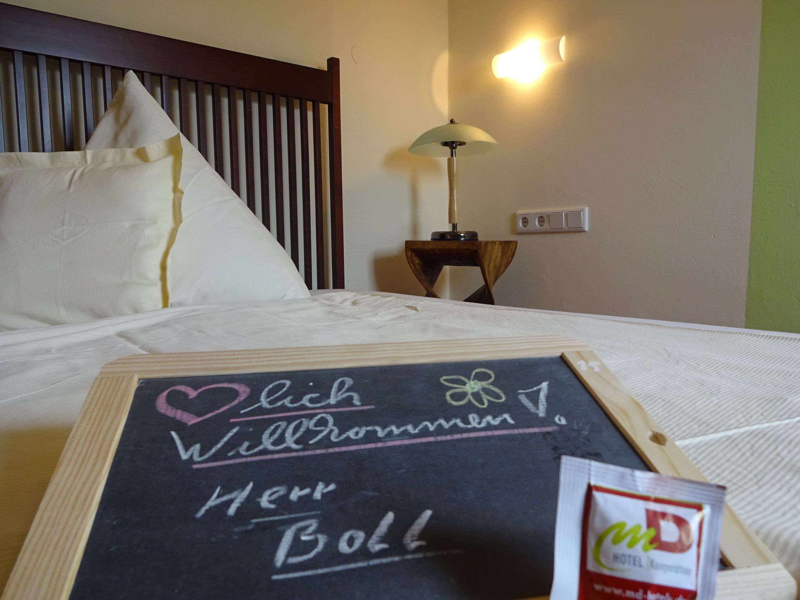 Una cama bien hecha con un cartel de bienvenida, una lámpara de noche y un enchufe en la pared.
