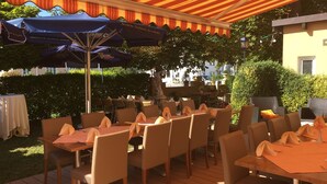 Un espace de restauration en plein air, avec des tables et des chaises, abrité par un auvent à rayures et entouré de verdure.