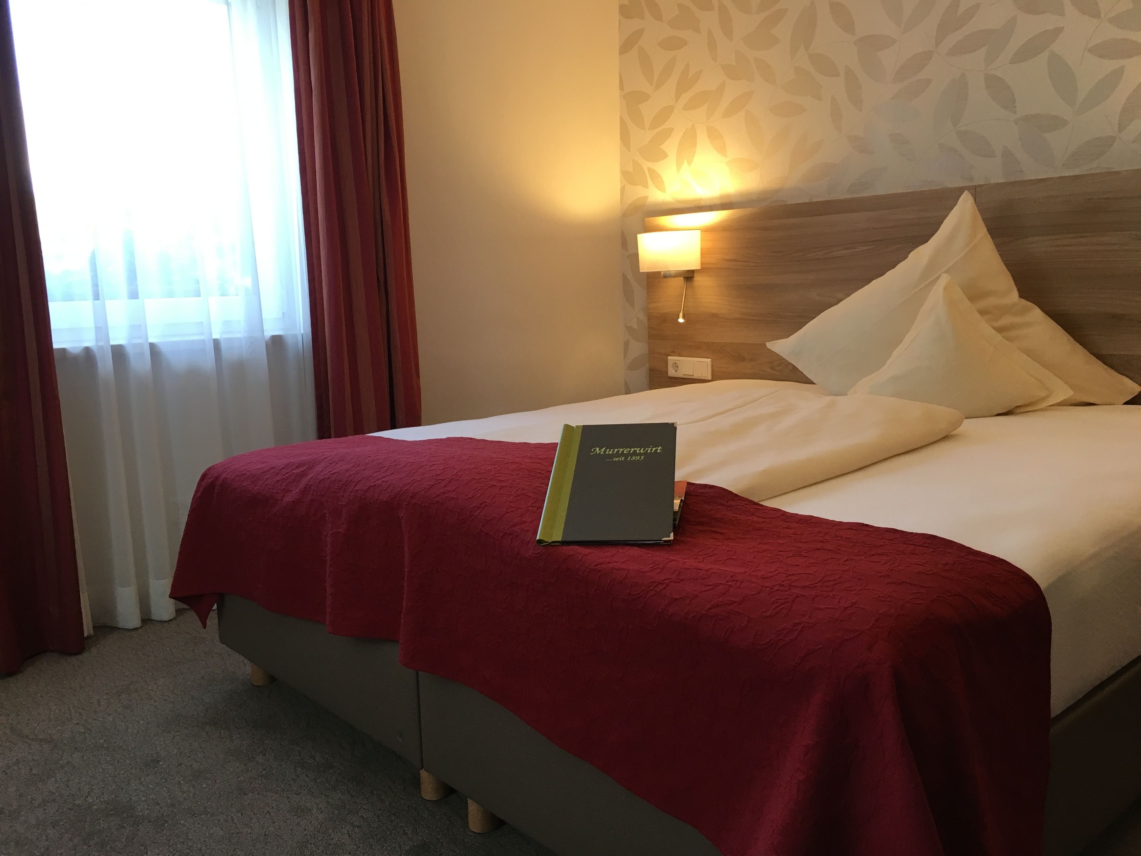 Una habitación de hotel con una cama, una colcha roja, un libro titulado 'Murrerwirt' y una mesita de noche con una lámpara.