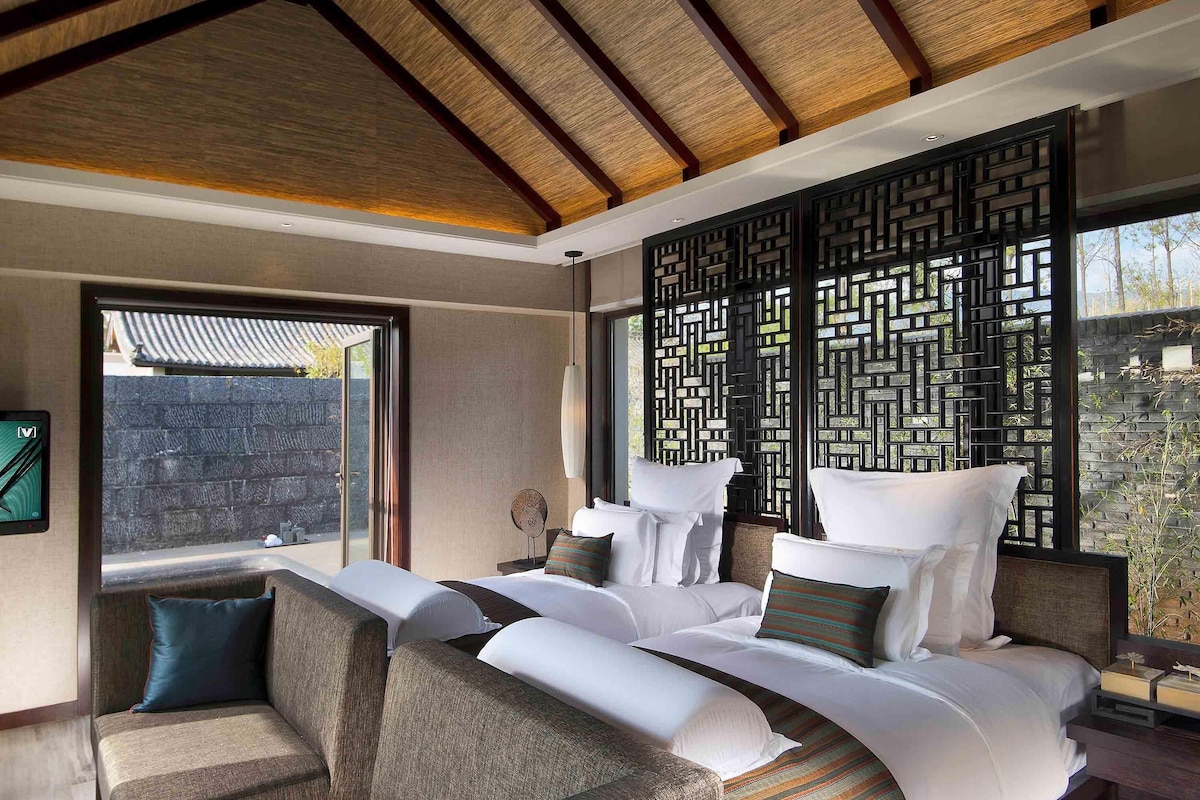 A modern hotel room with a large bed, a sofa, a decorative screen, and a view of the outdoors.