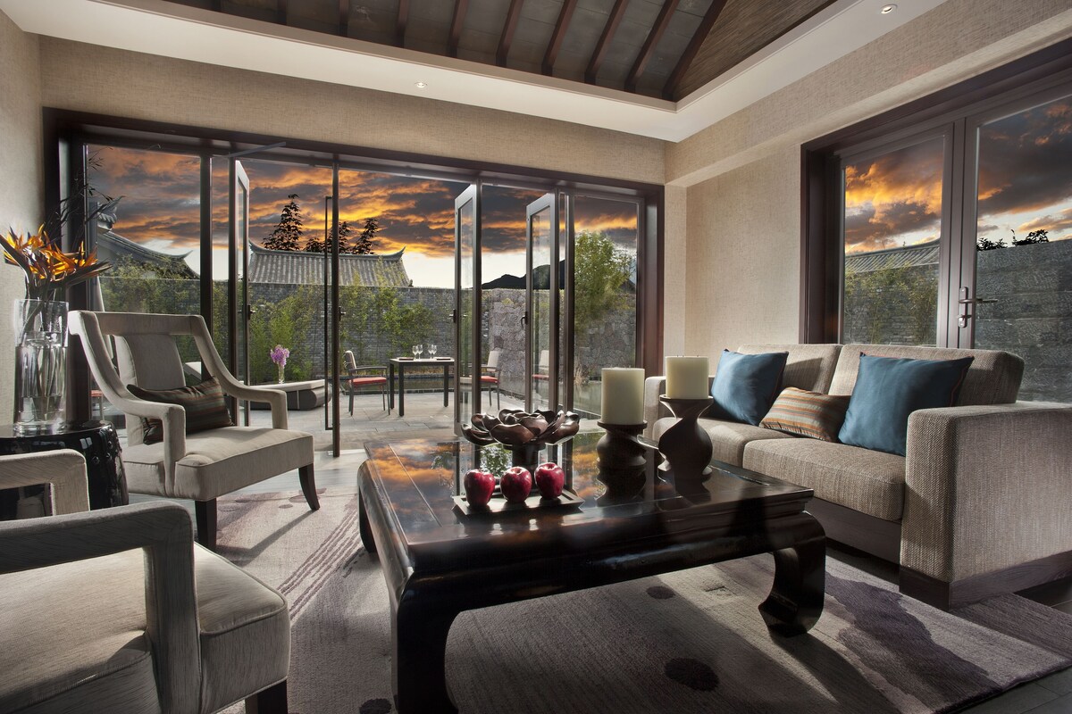 A modern living room with a sofa, chairs, and a coffee table. There is a large window with a view of traditional architecture and a sunset.