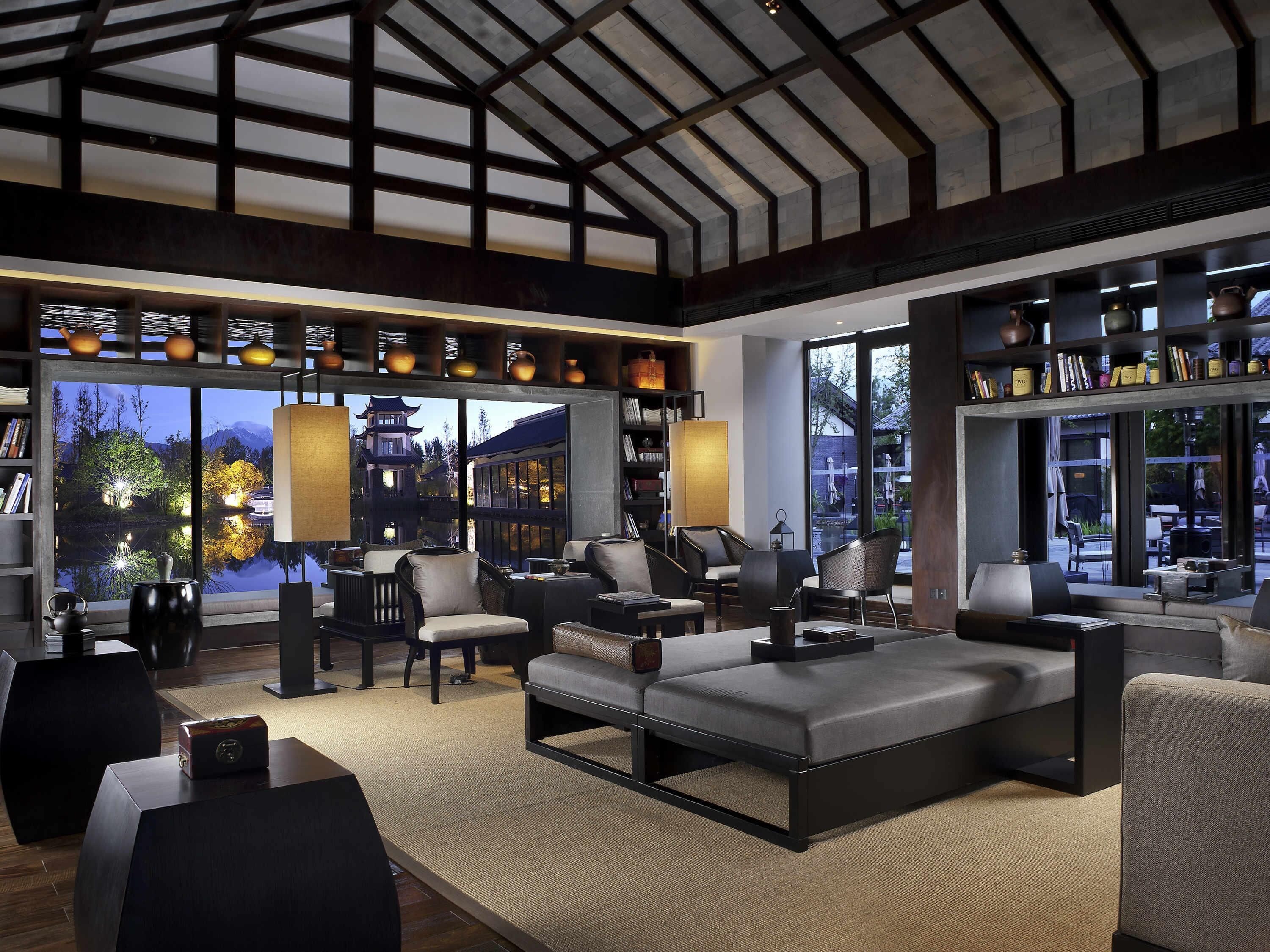 A spacious living area with a large sofa, multiple chairs, a coffee table, and a bookshelf. A large window offers a view of a traditional building and trees.