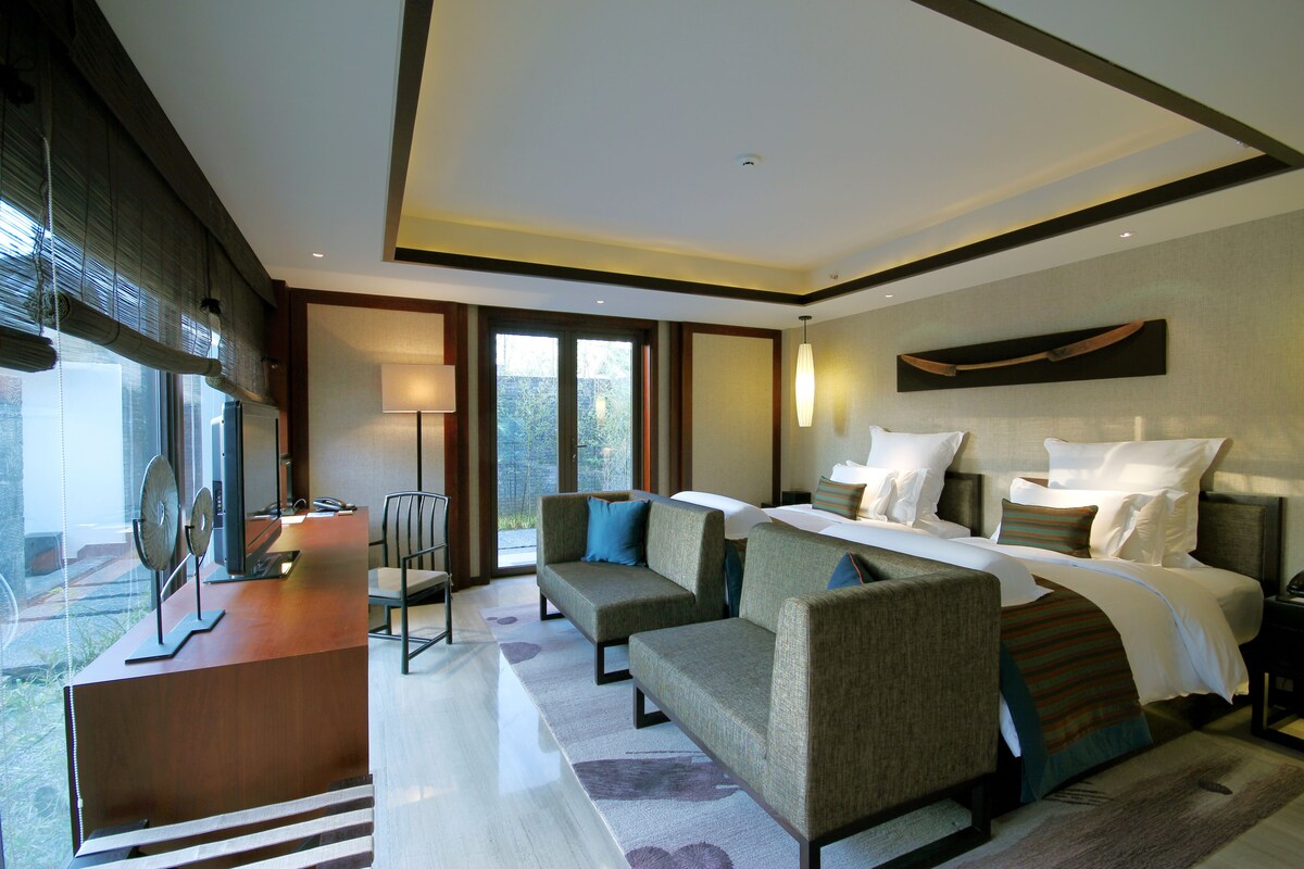A modern hotel room with a large bed, two sofas, a desk, and a view of the outdoors.