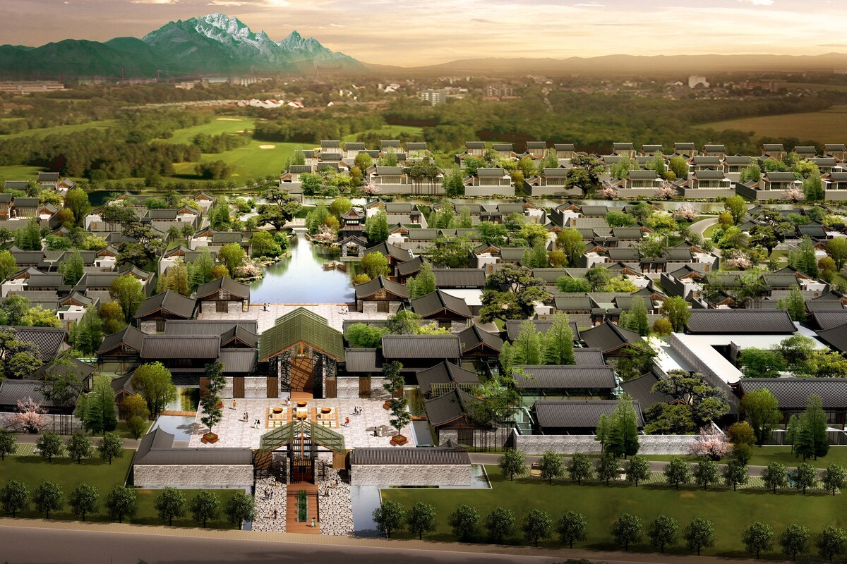 An aerial view of a traditional-style residential area with multiple houses, a central courtyard, and a mountain backdrop.