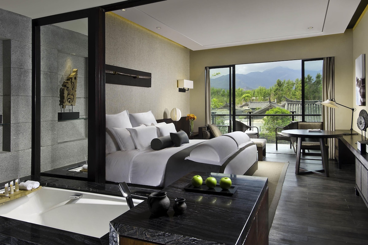 A modern hotel room with a large bed, a separate sitting area, a view of traditional architecture, and a bathroom with a bathtub.