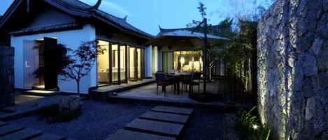 A traditional Japanese-style building with a tiled roof, a stone wall, and a gravel pathway leading to a seating area with tables and chairs.