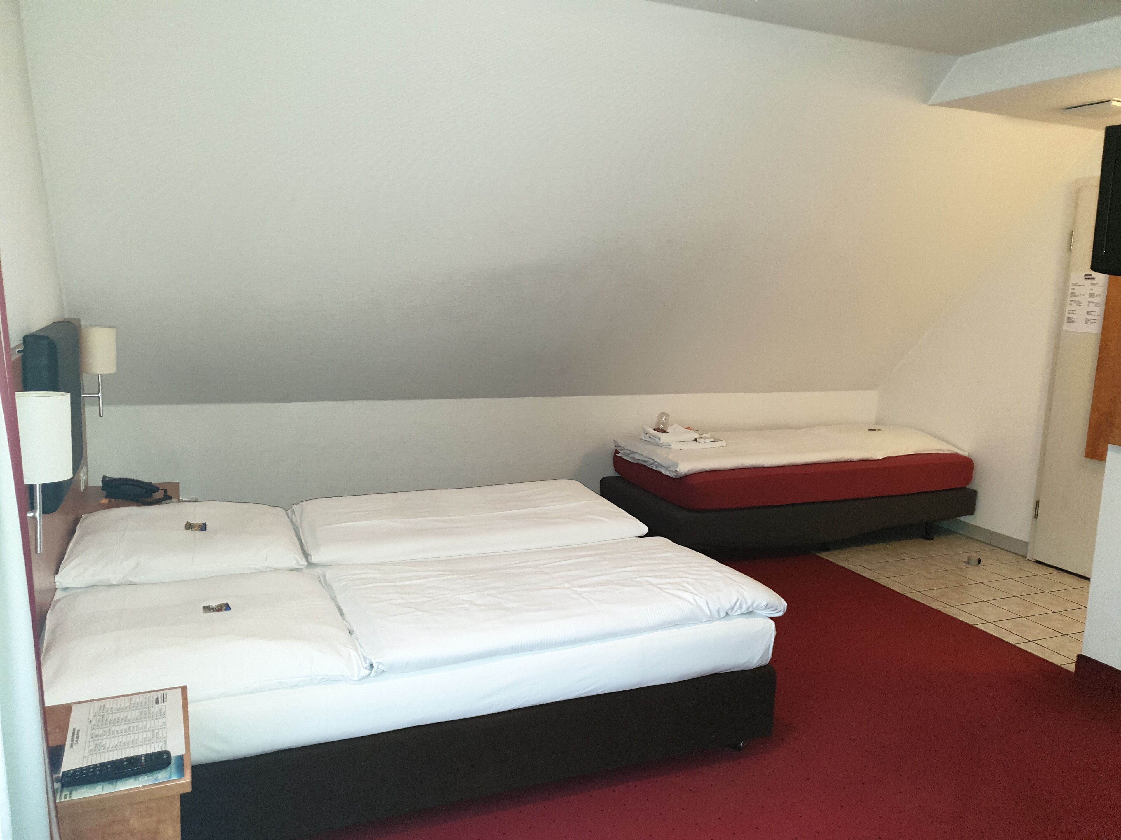A hotel room with two beds, a desk with a lamp, and a red carpet.